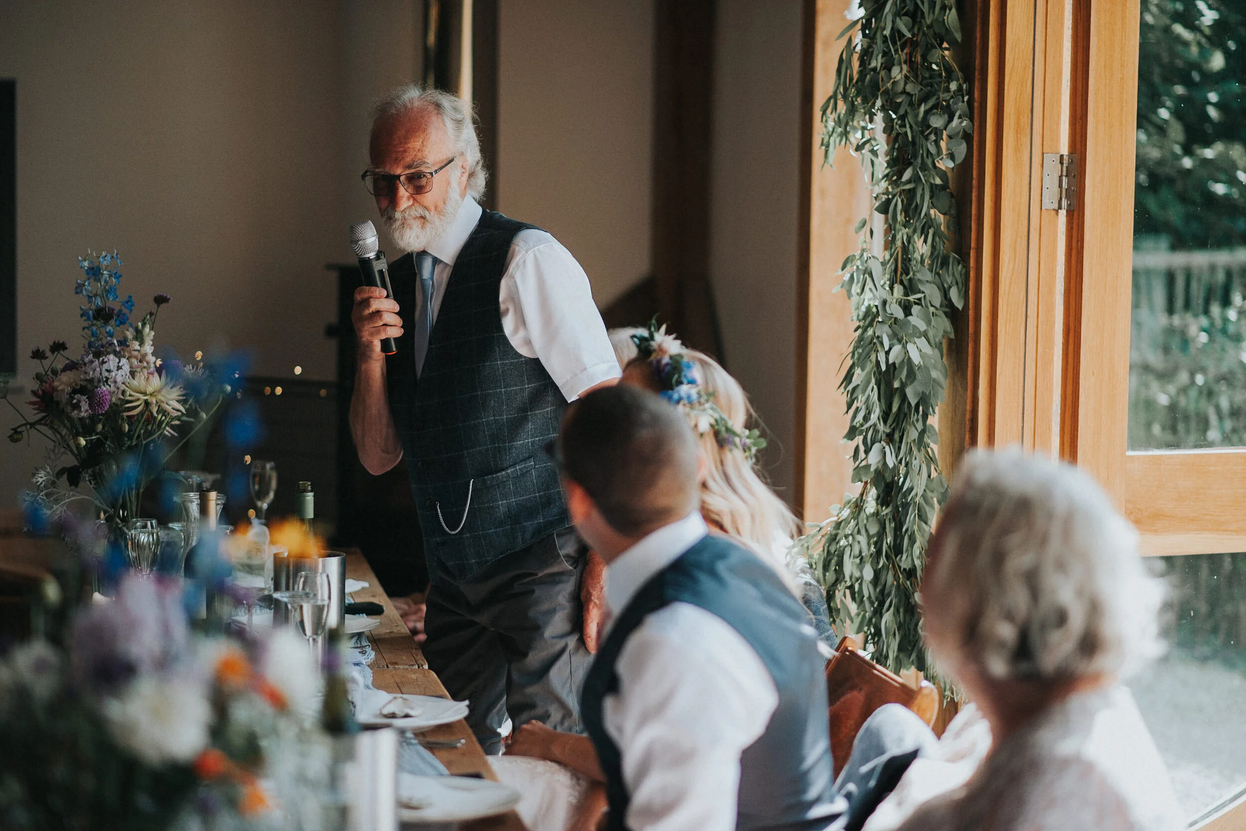 An elderly man with white hair and beard, wearing glasses, a white shirt, and a plaid vest, giving a speech with a microphone at a wedding reception. Guests are seated at a table decorated with flowers and glasses, listening to him. The setting appea