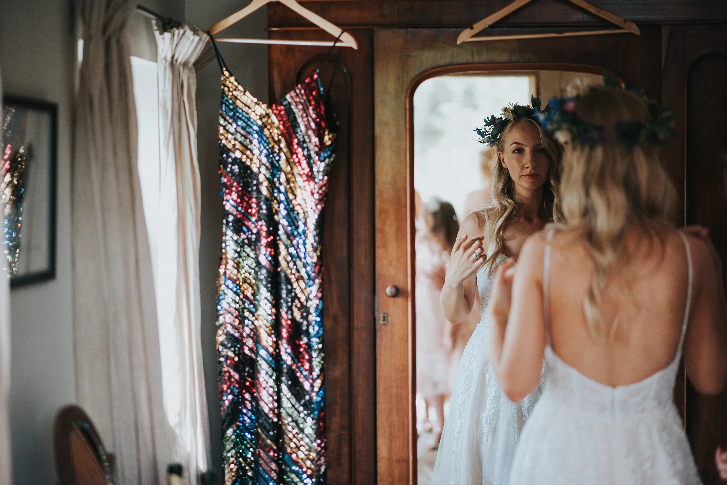 A bride with a floral crown looking at herself in a mirror, wearing a white wedding dress. A colorful, sequined dress is hanging near the mirror, and a curtain is partially open on the left side.