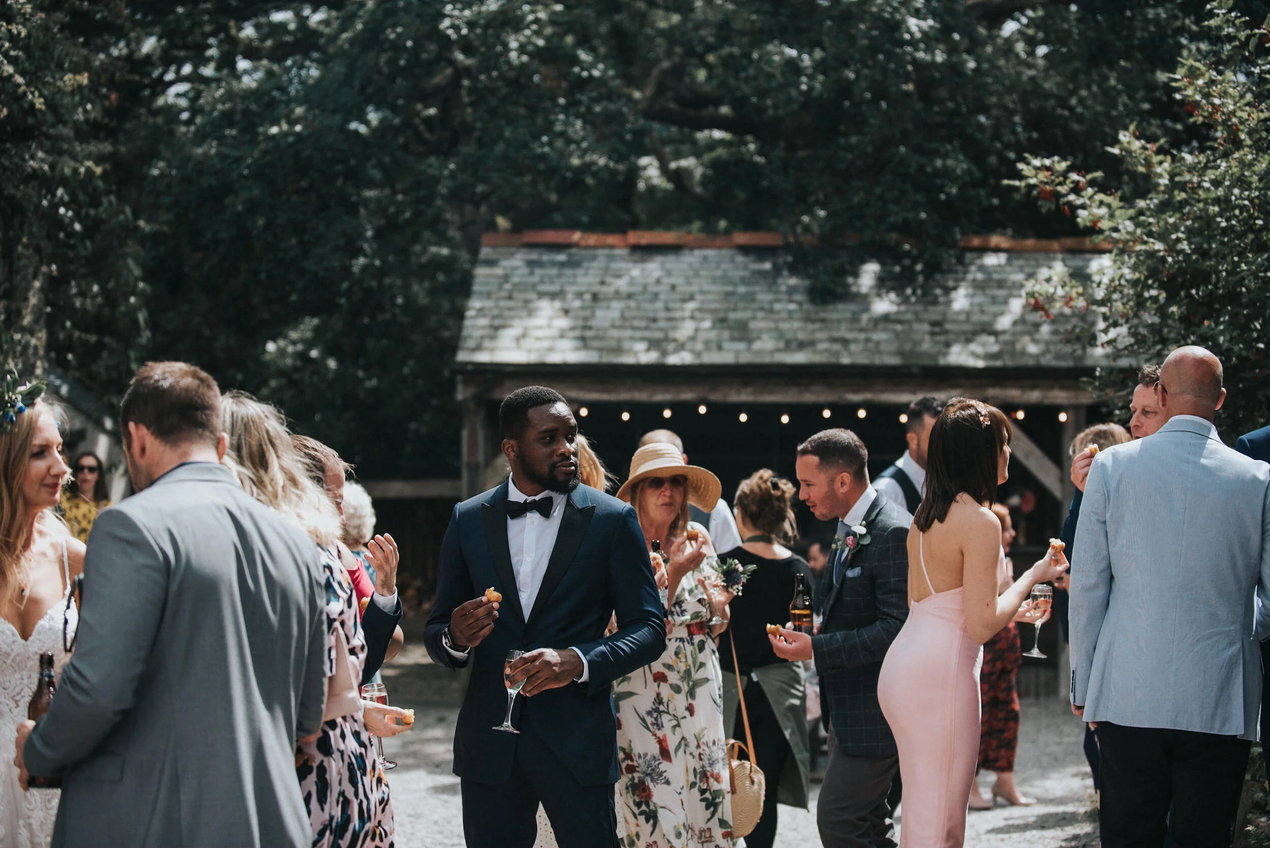 Group of people at an outdoor wedding reception, dressed in formal attire, holding drinks and chatting, with a rustic wooden structure and string lights in the background.