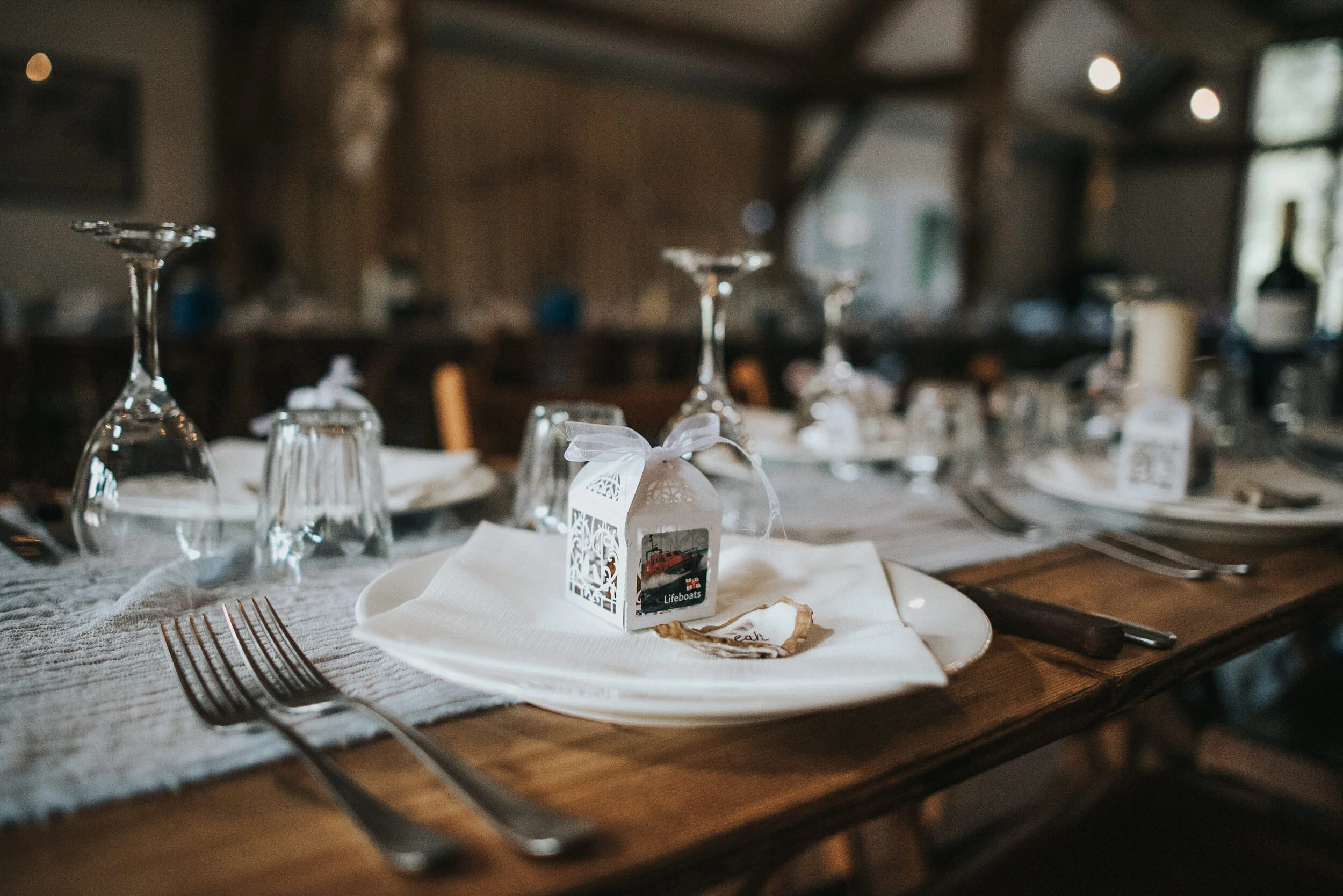 A rustic dining table set for a special occasion with wine glasses, plates, and silverware. There is a small white box with lace details and a ribbon, and a cookie on a napkin. The background shows a wooden interior with soft natural light.