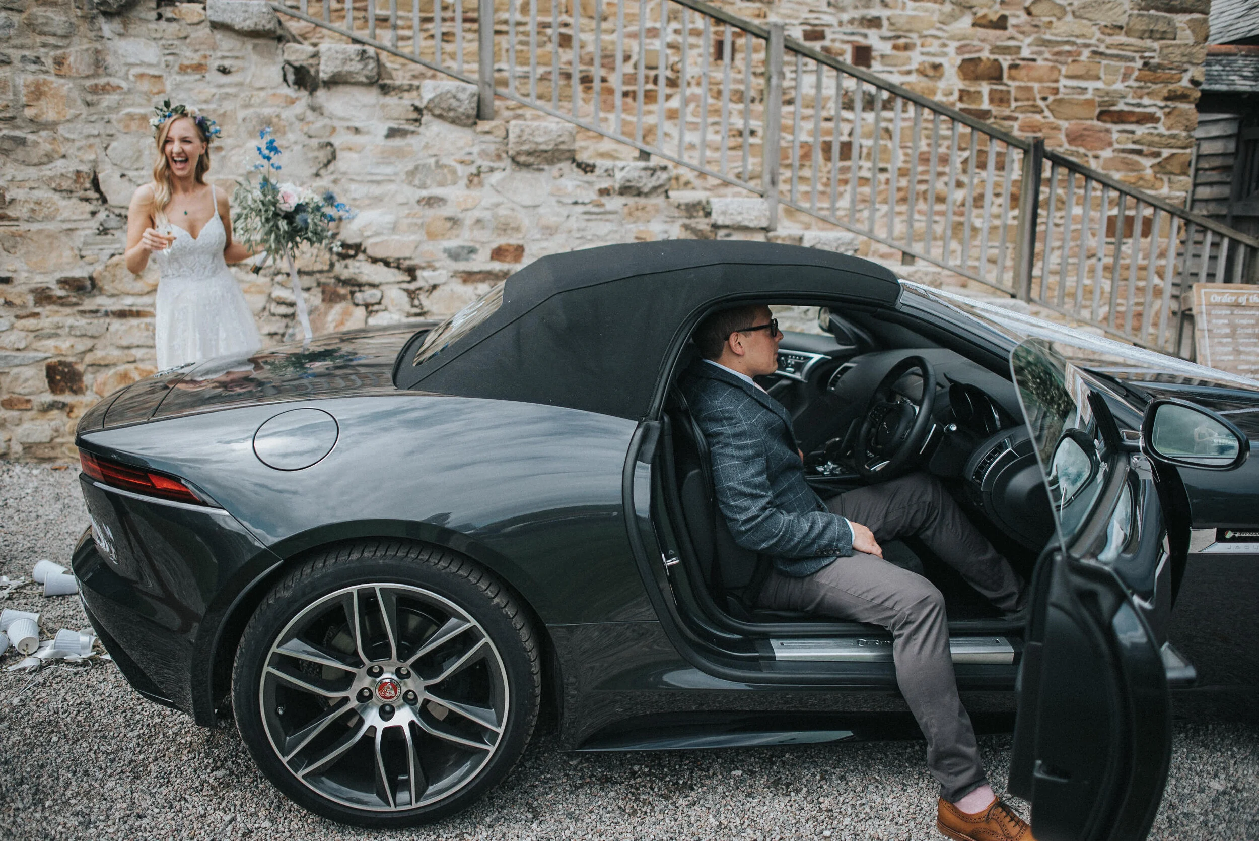 Nancarrow Farm Wedding Photography, groom in black sports convertible car with the door open. A woman in a white dress with a floral headband is standing outside the car, smiling and holding a bouquet of flowers.
