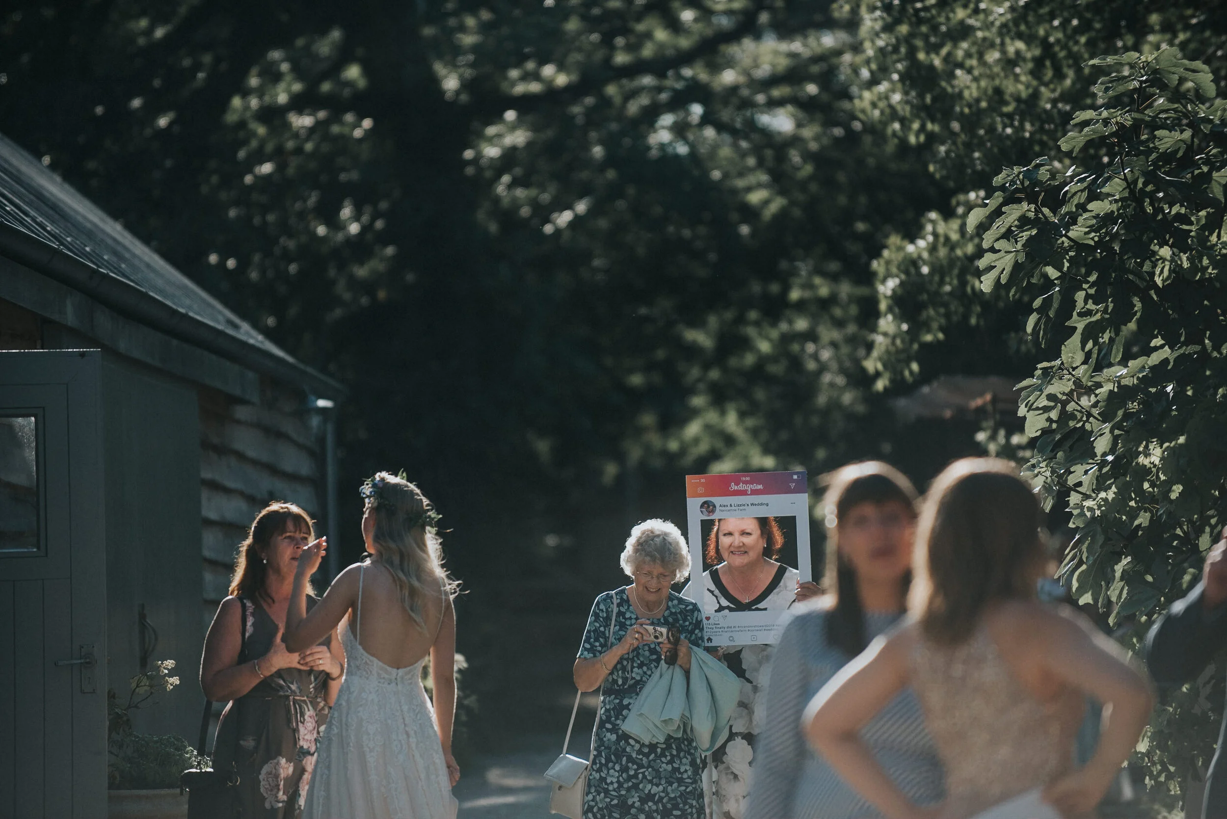 Group of women gathered outdoors, some taking photos, others engaging in conversation, with greenery and trees in the background.