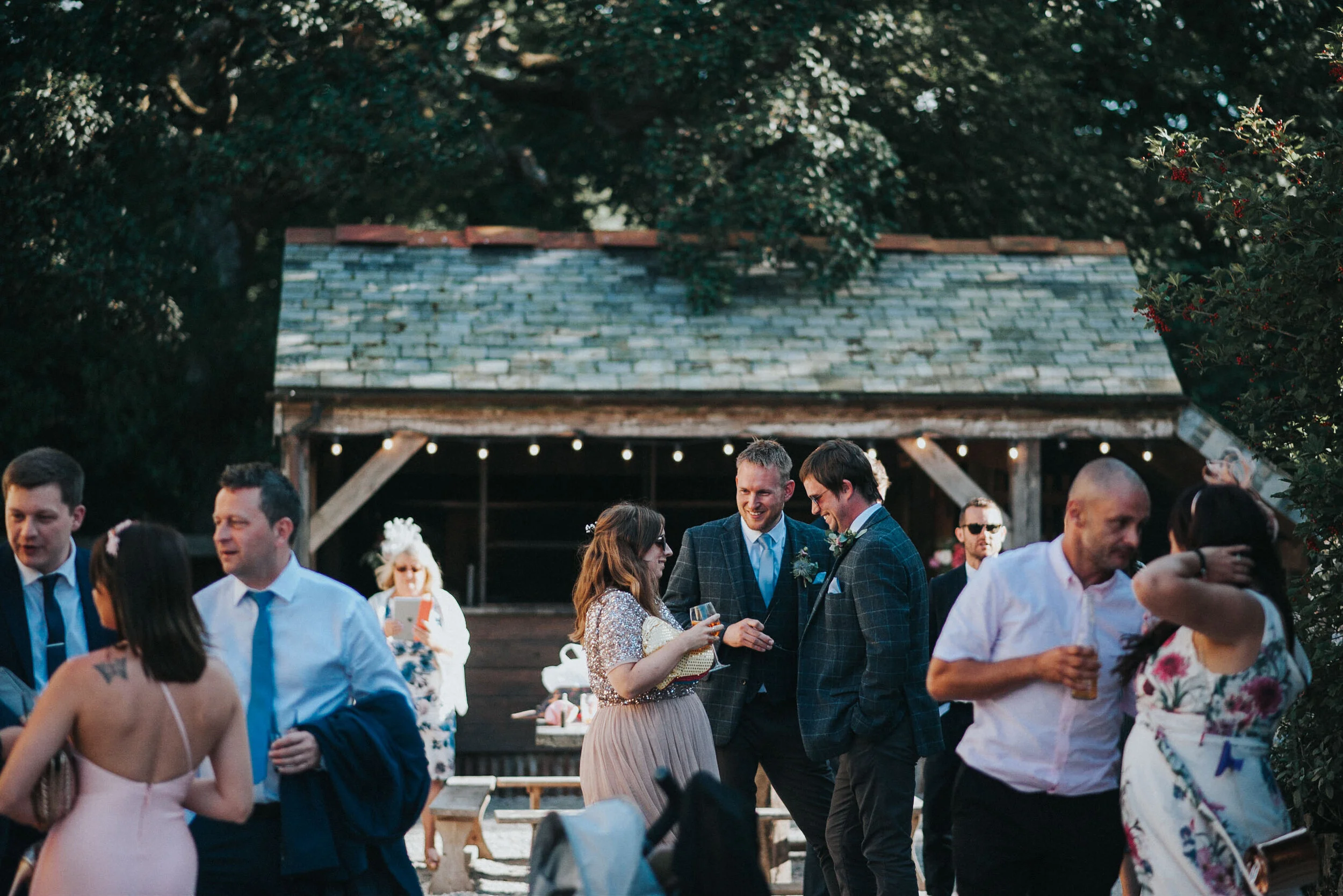 Group of people at outdoor wedding reception, some talking, some holding drinks, with a rustic wooden building and string lights in the background.