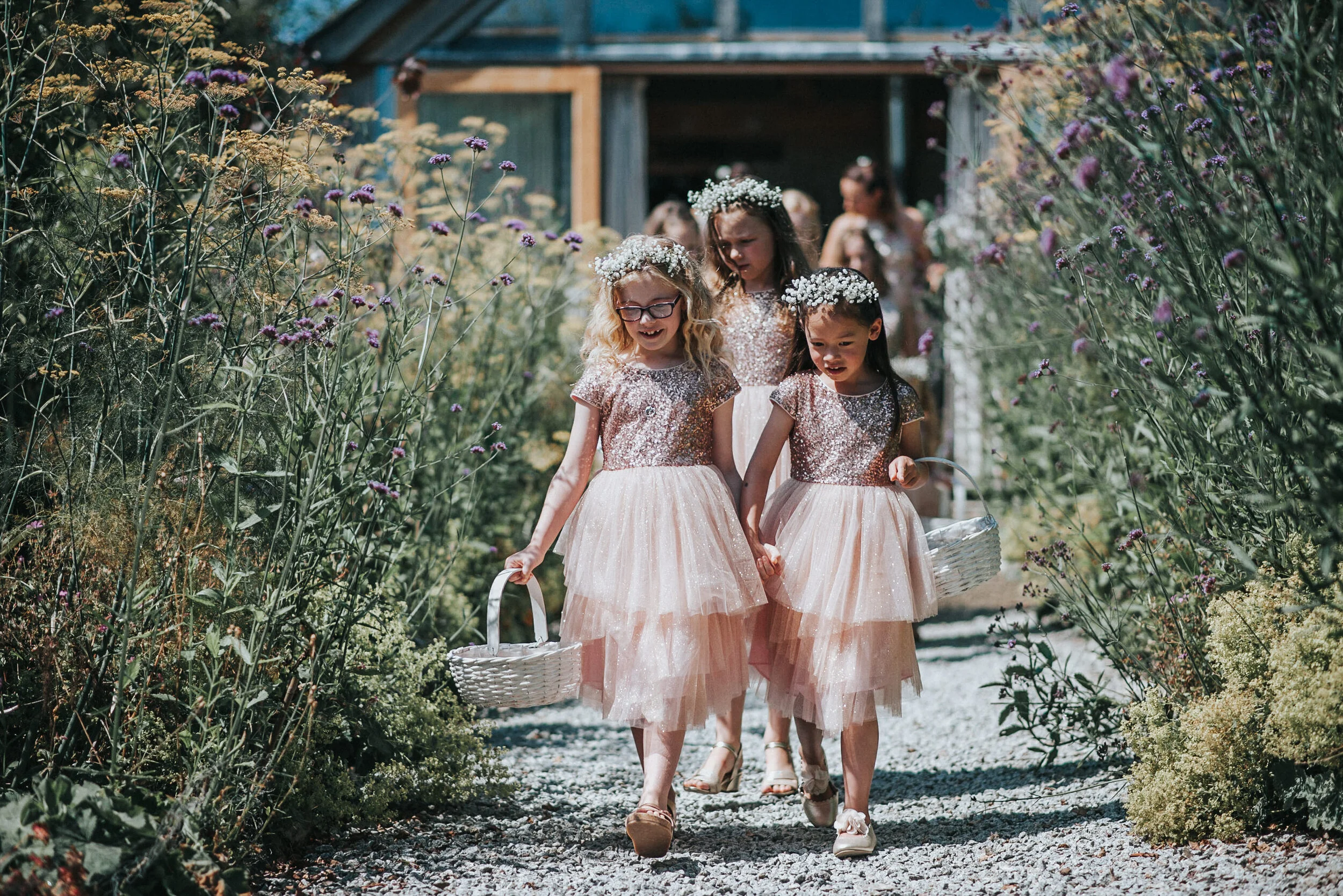 Three young girls in matching pink dresses with sequined tops and tulle skirts, carrying small baskets, walking down a gravel path surrounded by tall purple and cream flowers, with a wooden building in the background, during a sunny day.