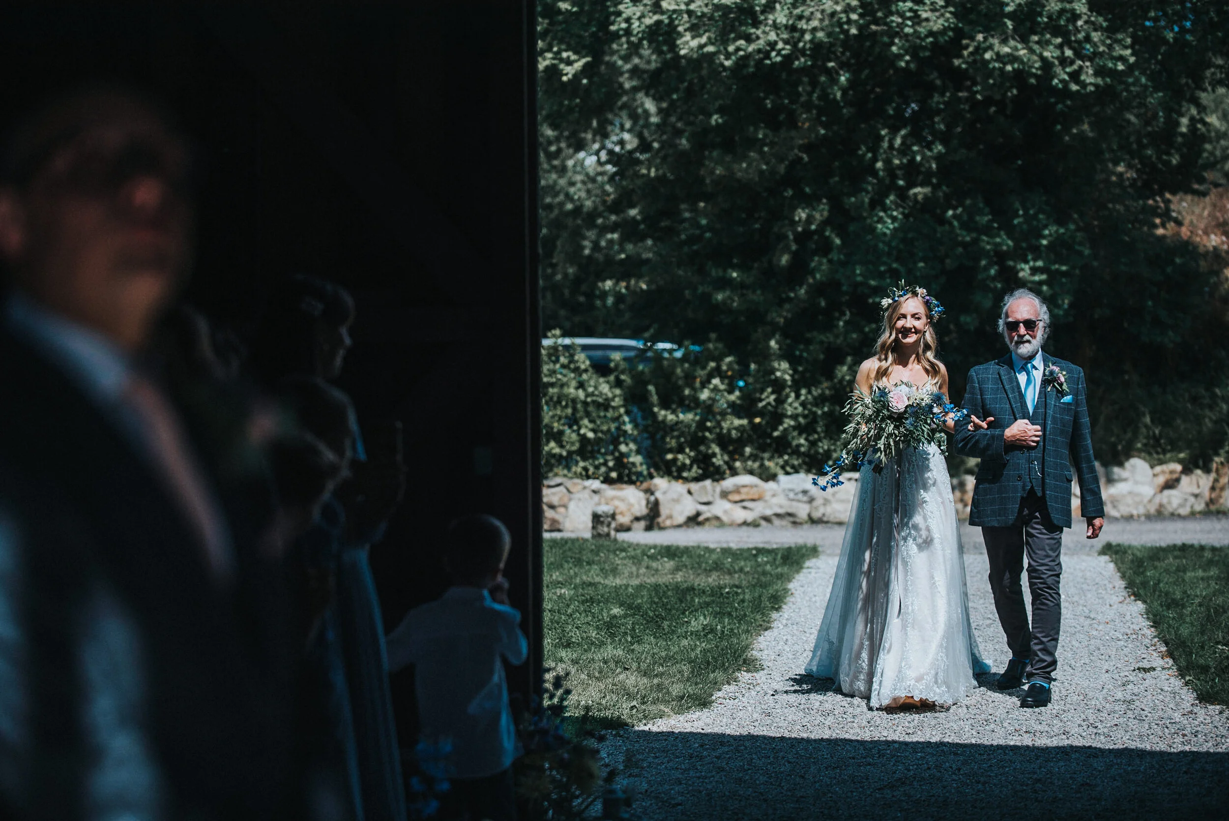 A bride walking down the aisle with an older man during a wedding ceremony outdoors, with guests visible on the left side of the image.