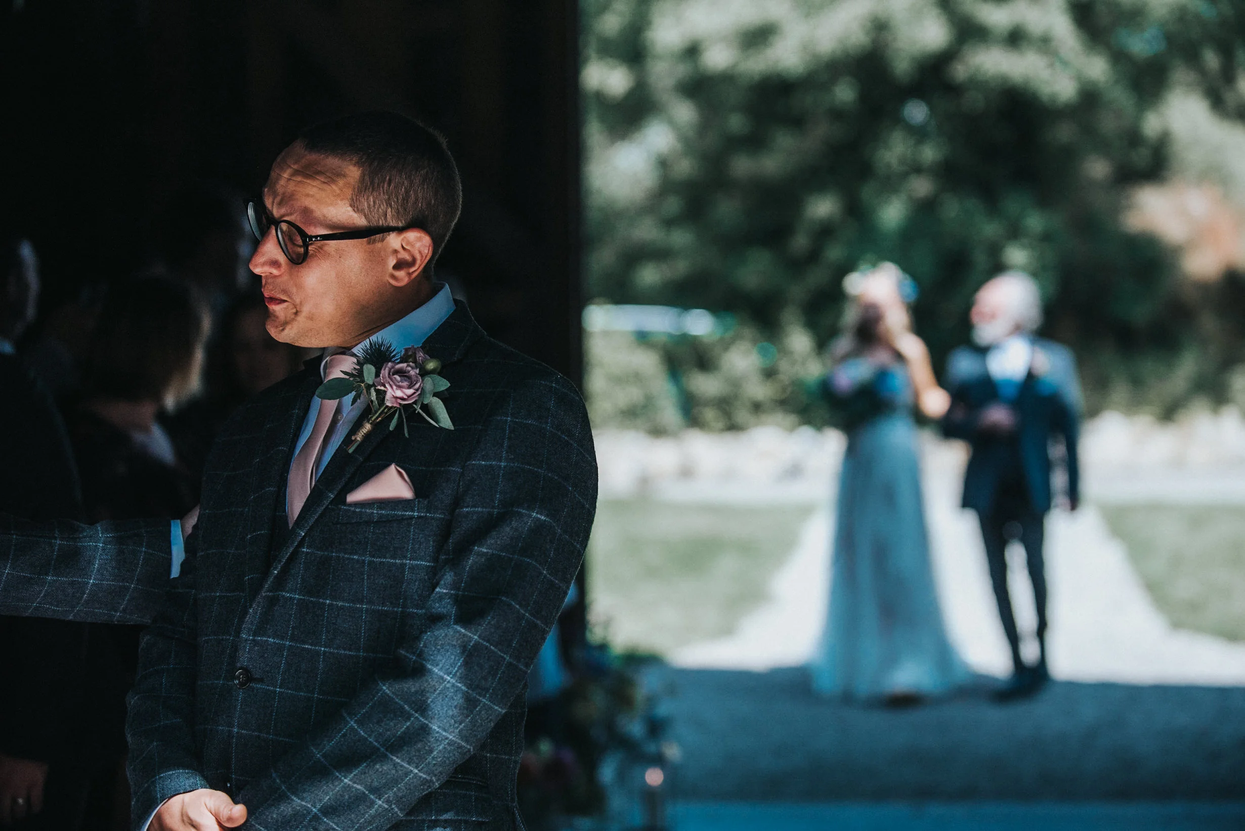 A man in a plaid suit with glasses, a pink boutonniere, and a pink pocket square standing indoors, looking down with a thoughtful expression. In the background, a bride and groom are walking outside along a pathway, blurred in the distance.
