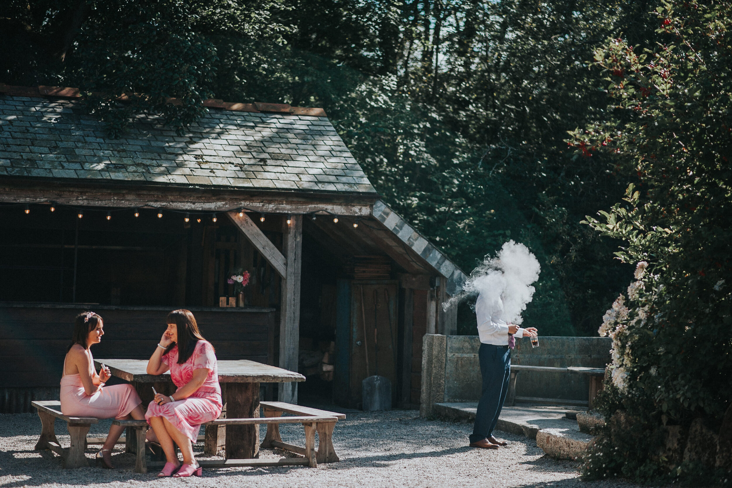 Two women are sitting at a rustic outdoor table, having a conversation, while a man nearby exhales vapor from an e-cigarette or vape device, with a wooden structure and trees in the background.