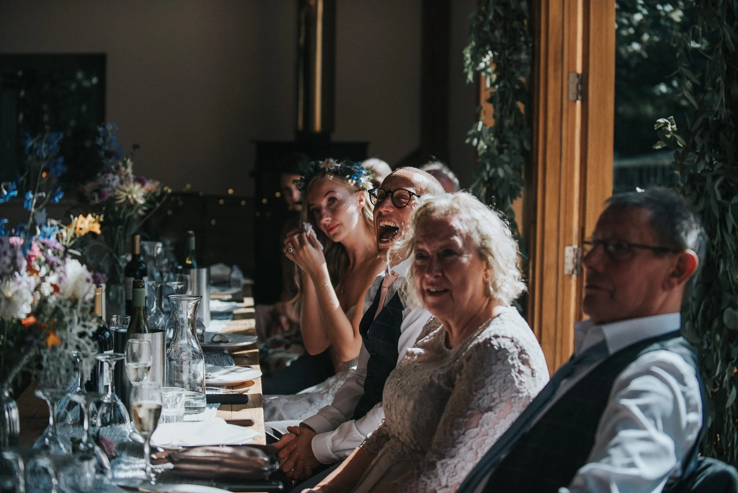 People sitting at a long table during a wedding reception, smiling and enjoying the moment. The table is decorated with flowers and glasses, set for the celebration.