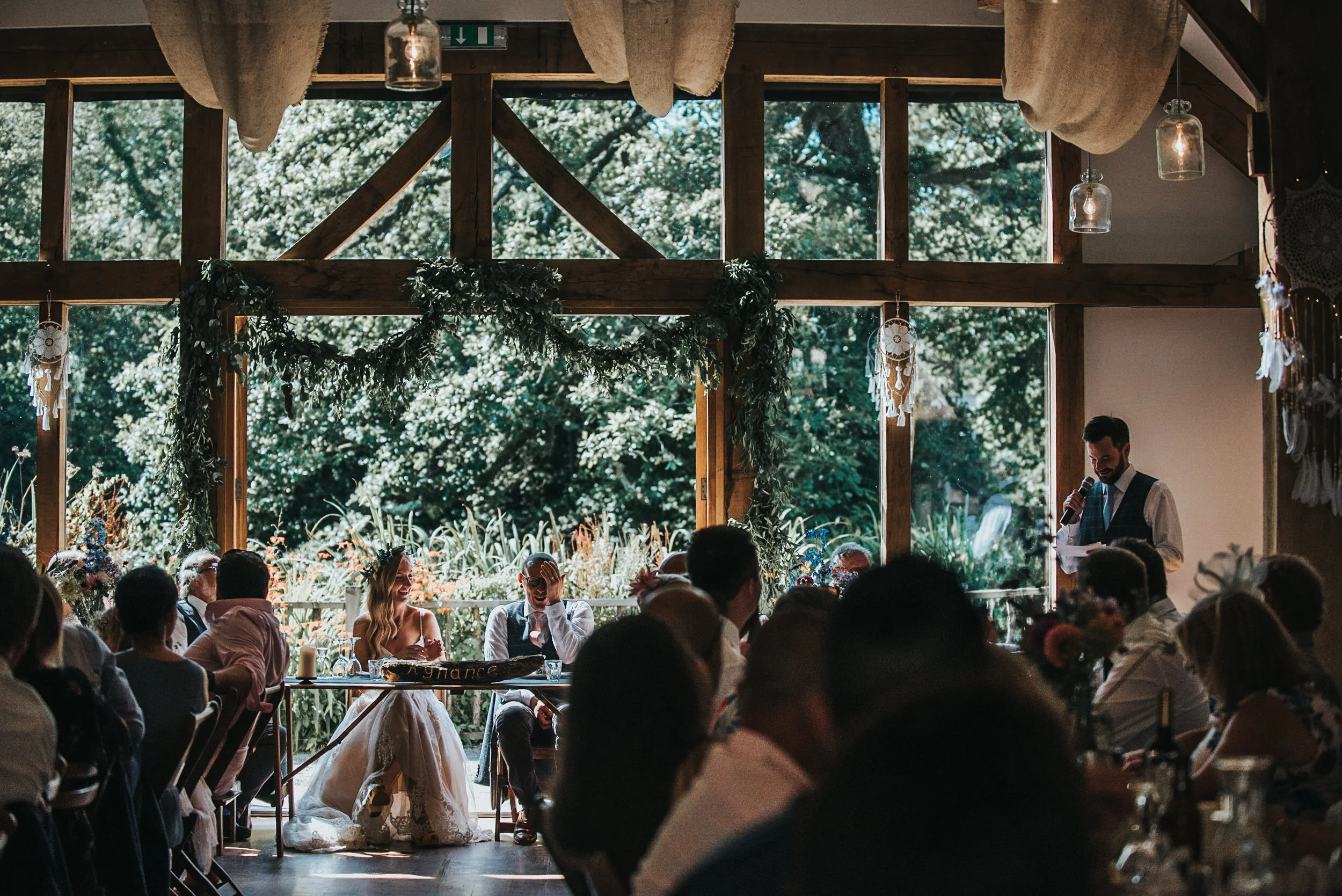 A wedding reception inside a rustic wooden venue with large windows, decorated with greenery and dreamcatchers, where a bride and groom sit at a table while a man in a vest gives a speech.