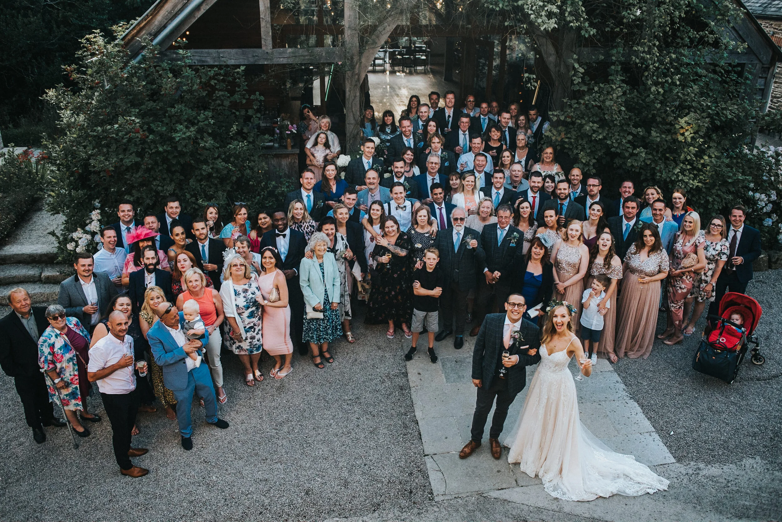 A large group of wedding guests gathered outside a rustic venue, with a bride and groom in the foreground smiling and holding drinks.