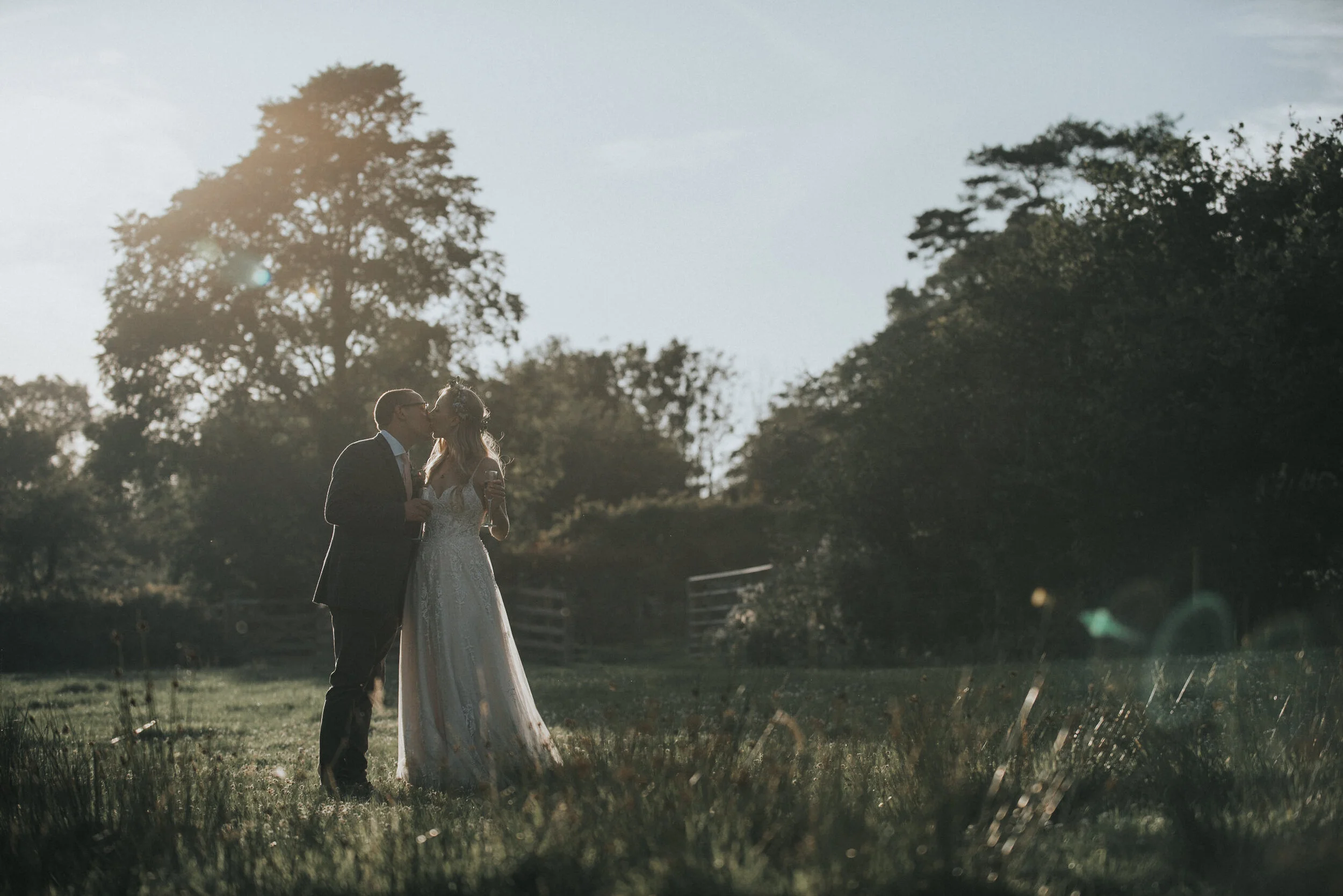 A bride and groom sharing a kiss outdoors during sunset, standing on grass with trees and a wooden fence in the background.