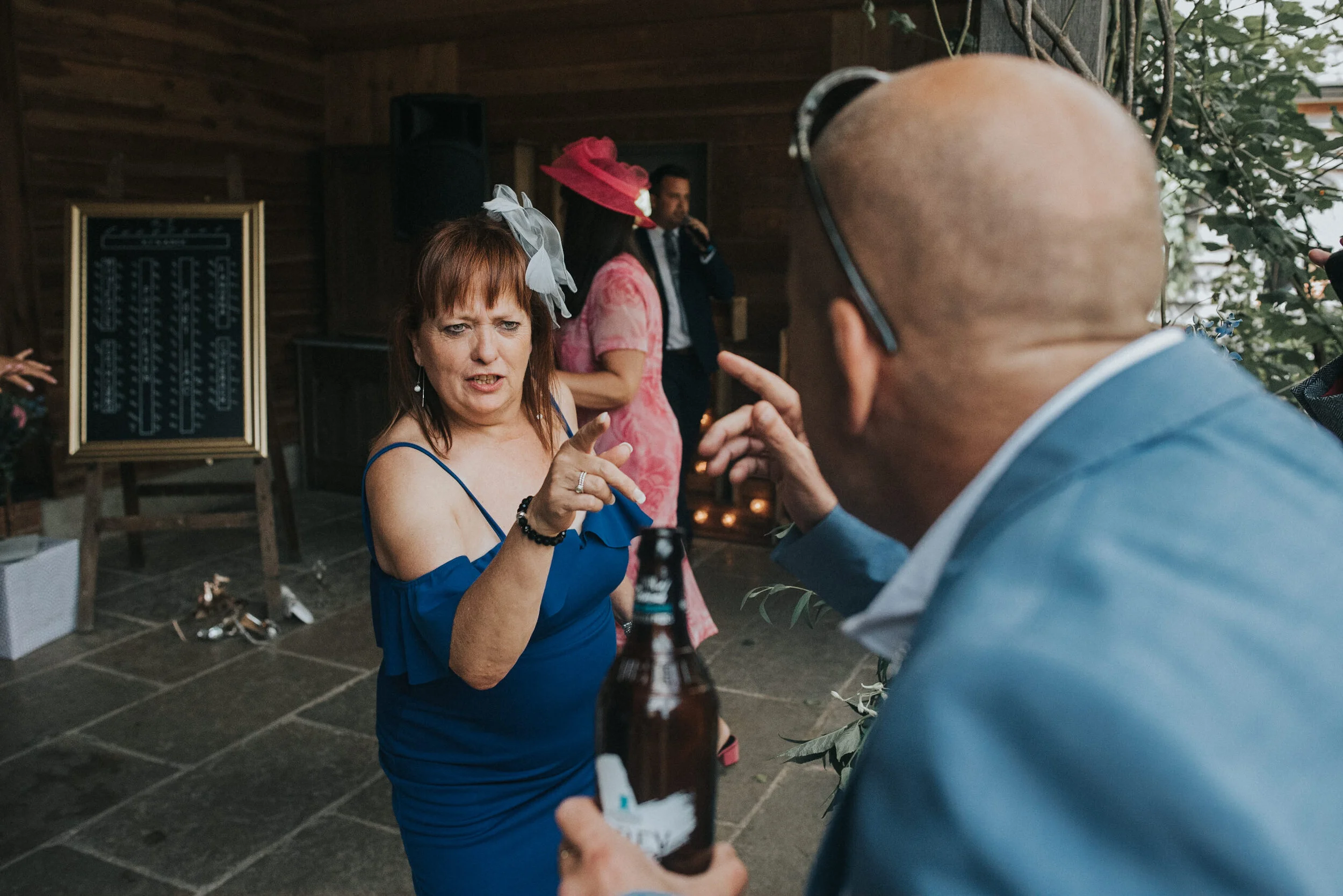 A woman in a blue dress with a large gray bow in her hair appears to be arguing with a bald man in glasses who is holding a beer bottle. The woman is pointing her finger at the man. In the background, there are other people, including a woman in a pi
