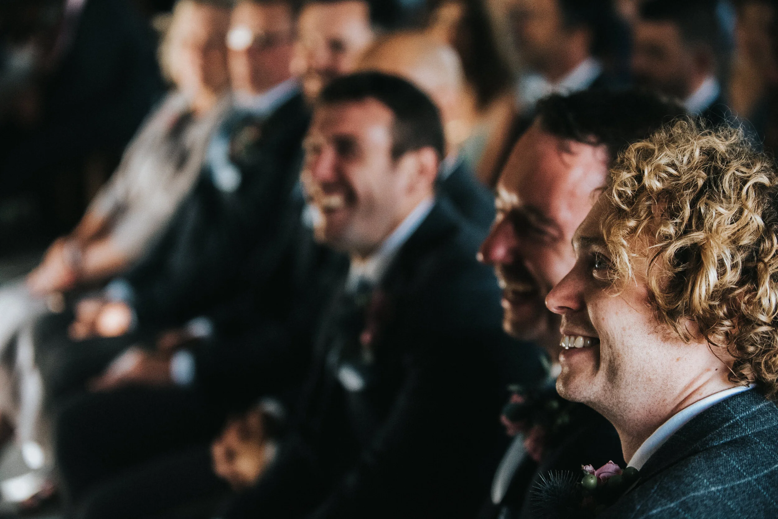 Group of men dressed in suits, smiling and laughing during a formal event, with focus on a curly-haired man in the foreground.