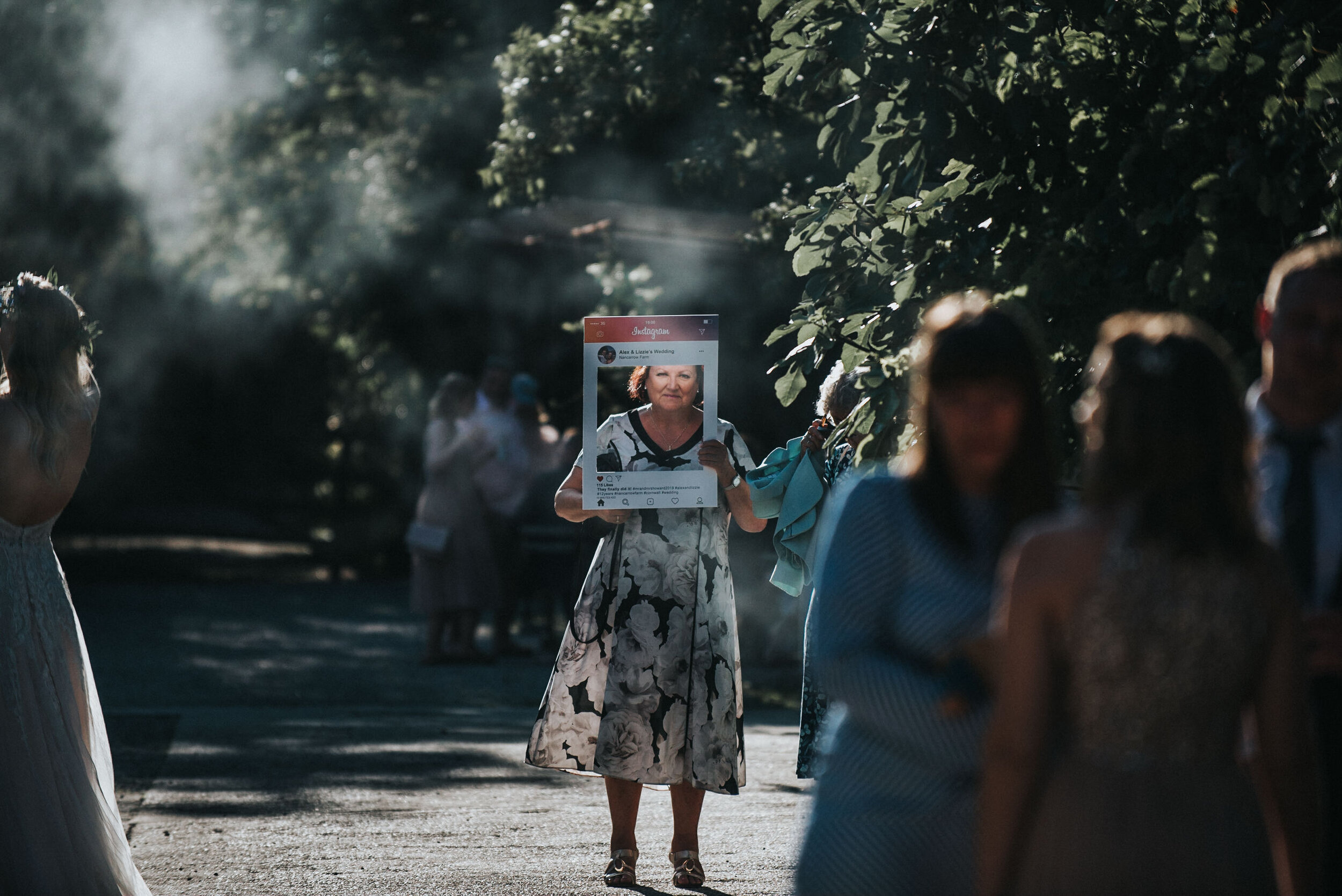 A woman holding a photo frame at a social gathering outdoors, surrounded by other people, with trees and sunlight in the background.
