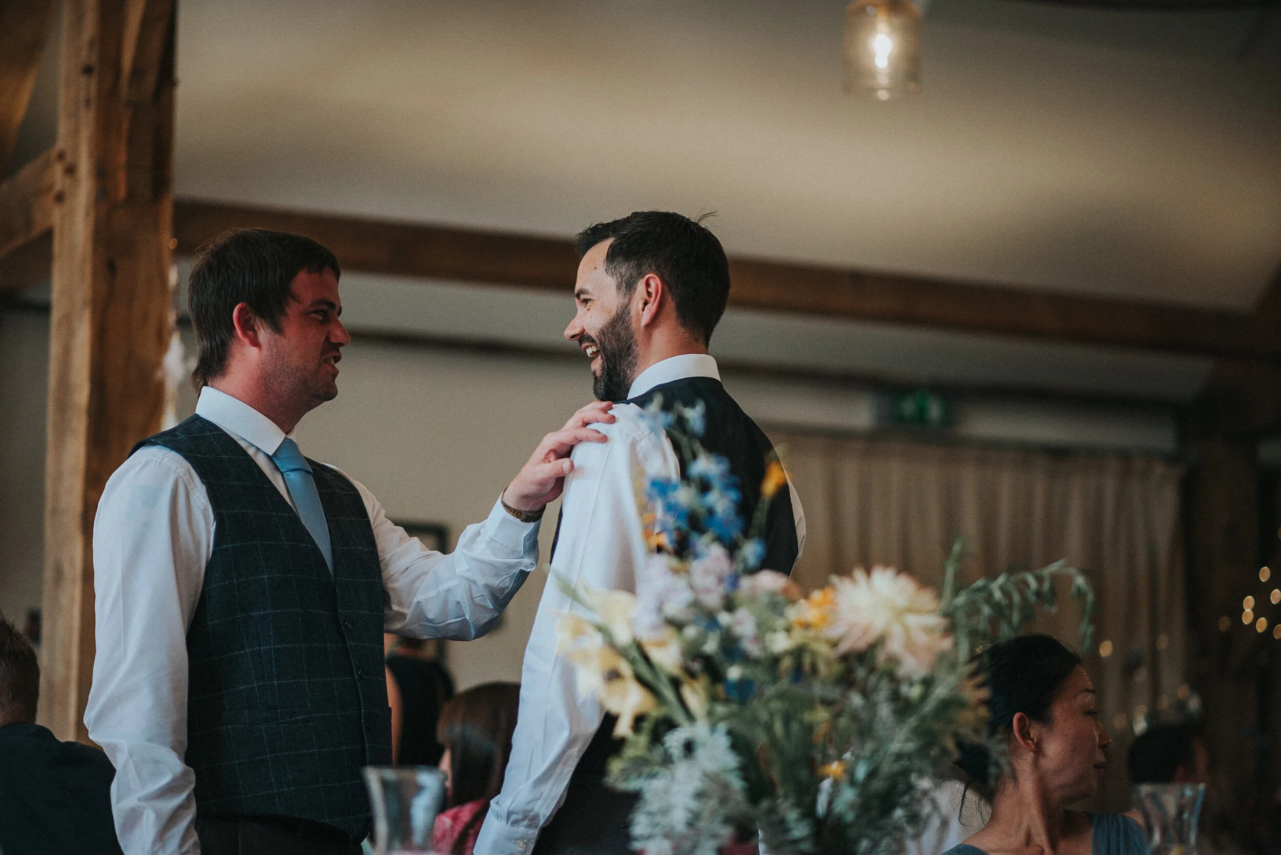 Two men with dark hair and beards smiling and interacting at a social event, one touching the other's shoulder, surrounded by floral arrangements and seated guests.