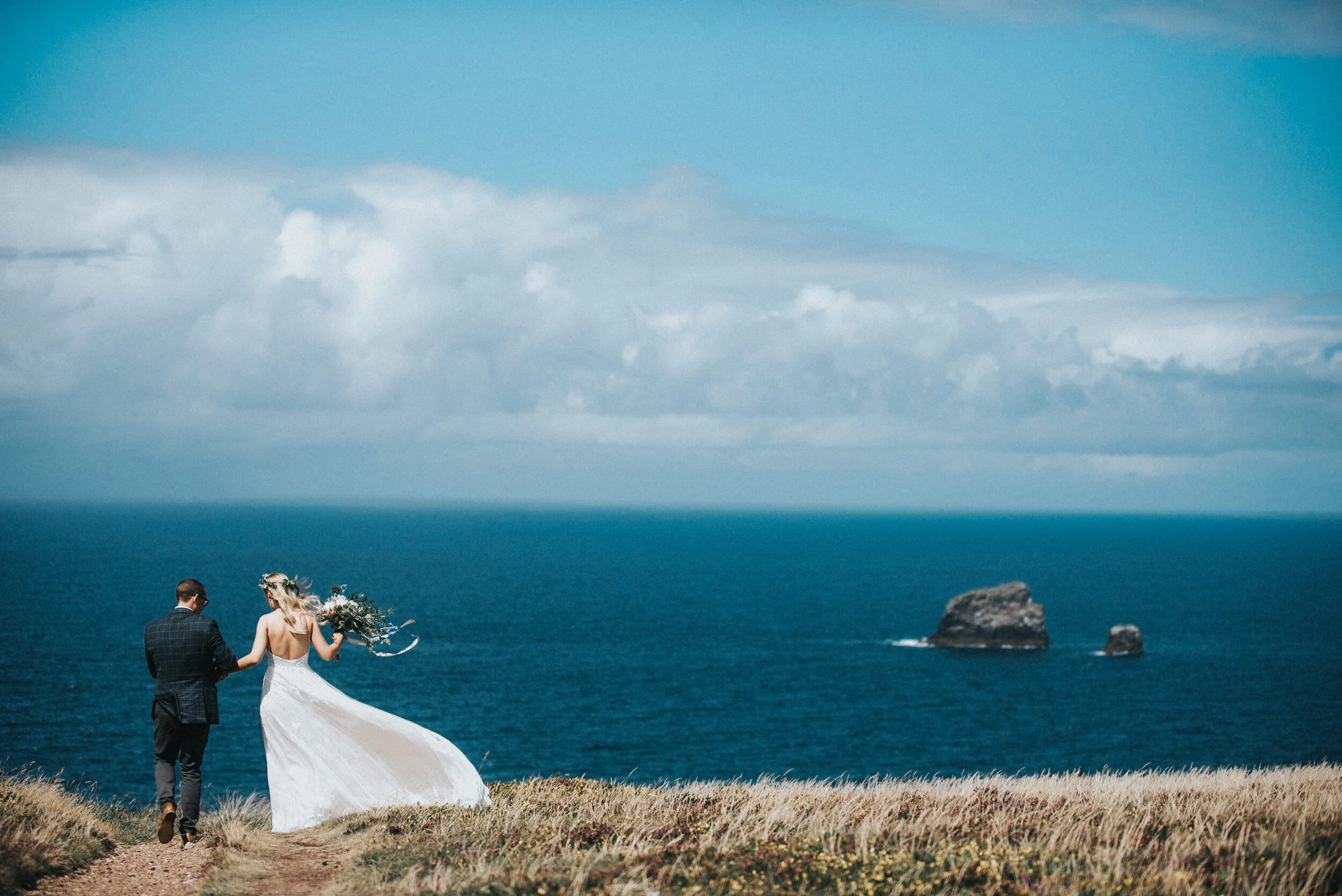 Bride and groom walking hand in hand along a grassy cliff overlooking the ocean on North coast of Cornwall with rocks in the water under a partly cloudy sky.