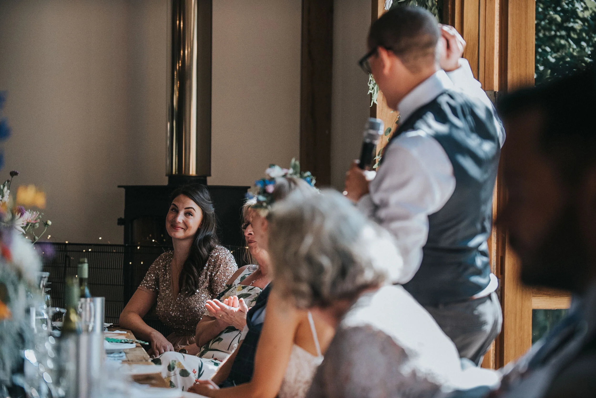 A man with glasses and a vest is giving a speech while holding a microphone, with a group of women seated at a table listening, in a warmly lit room with wooden accents.