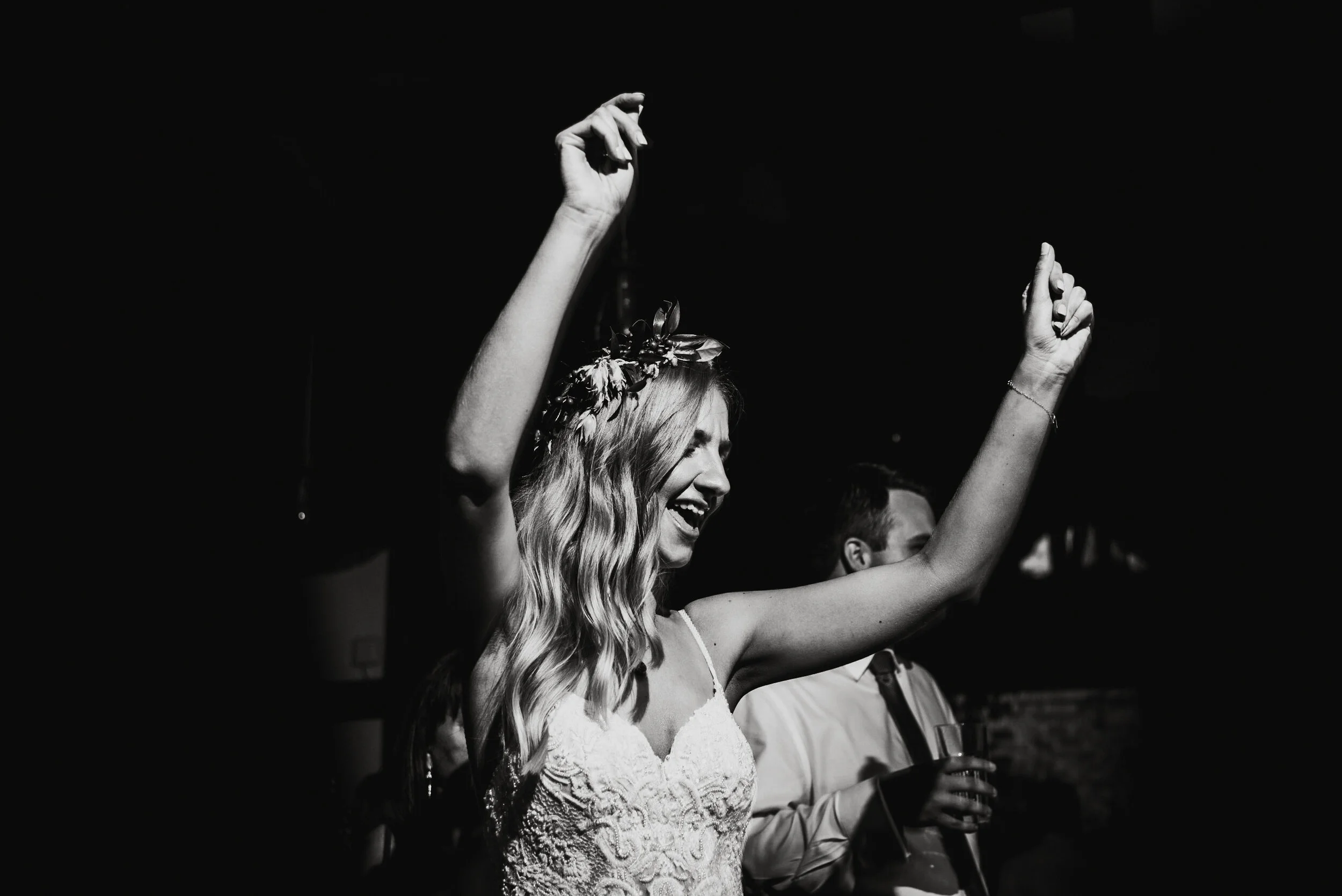 A woman with long wavy hair, wearing a lace dress and a floral headpiece, smiling and raising her hands in celebration at an event.