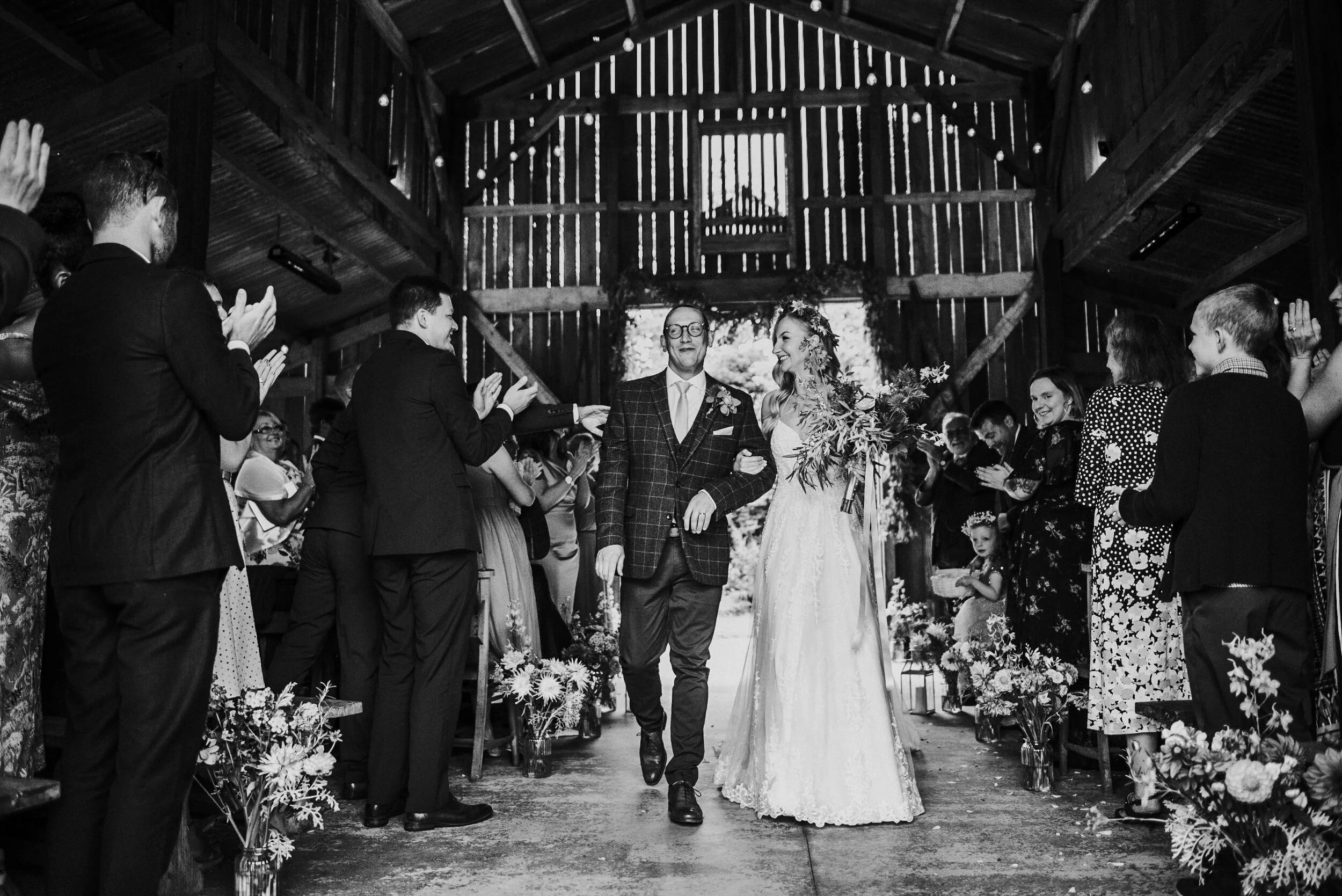 Black and white photo of a bride walking down the aisle in a barn, accompanied by her father, with guests clapping on either side.