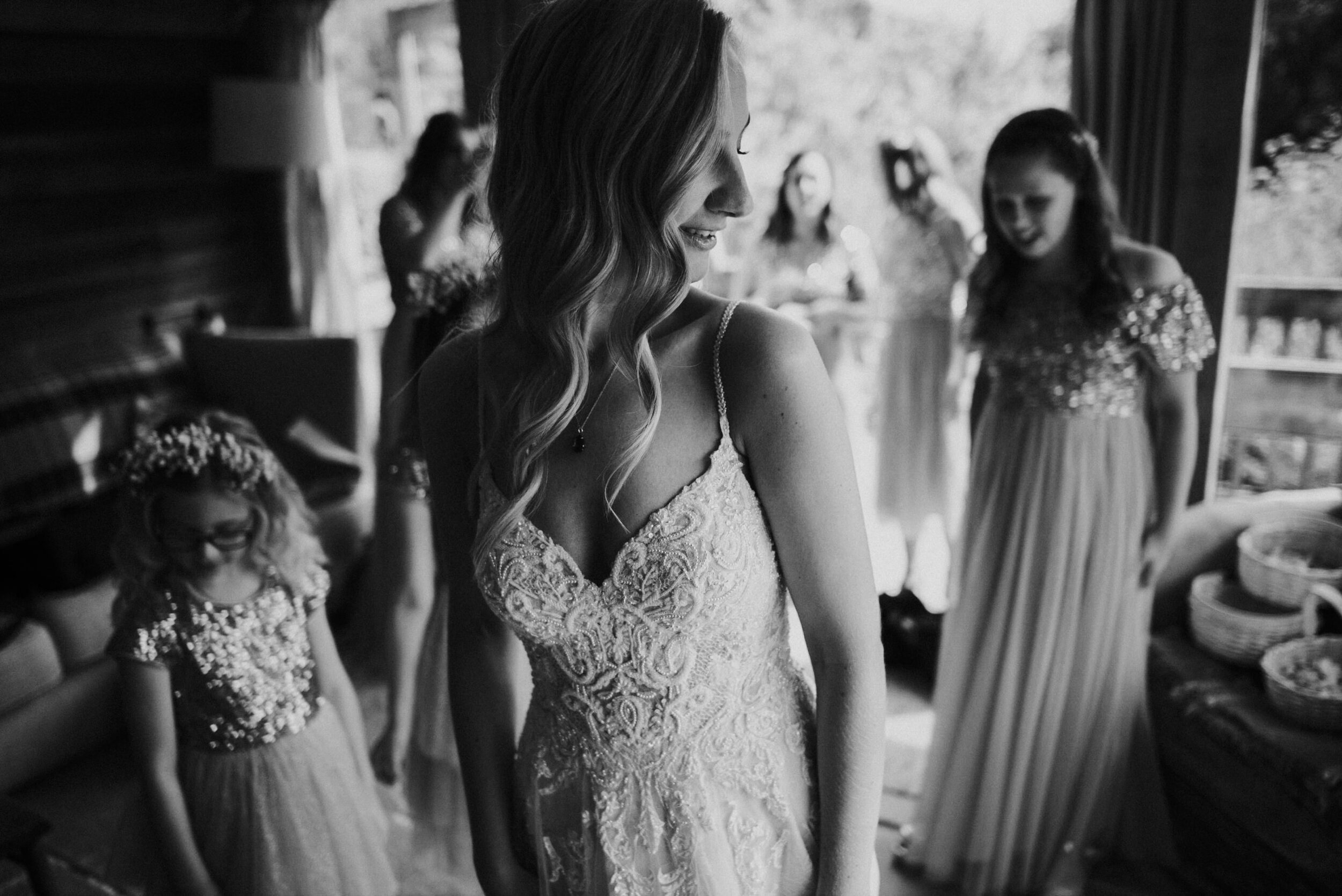A bride in a lace wedding dress with her eyes closed, smiling, standing in a room with bridesmaids and a young girl dressed in matching dresses, all preparing for a wedding.