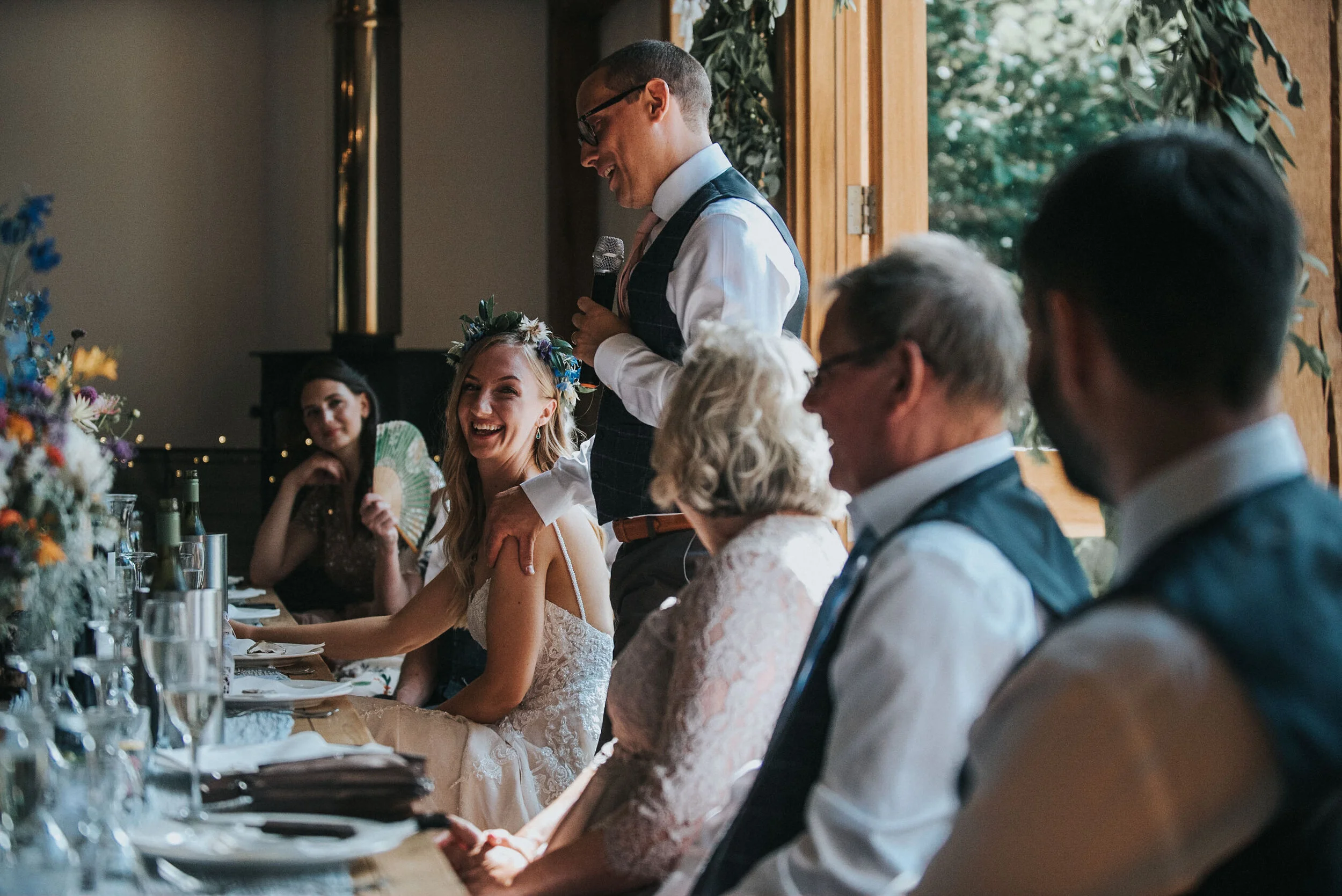 A wedding reception with a man giving a speech, standing next to a smiling bride wearing a floral crown, while guests seated at the table listen and enjoy the moment.