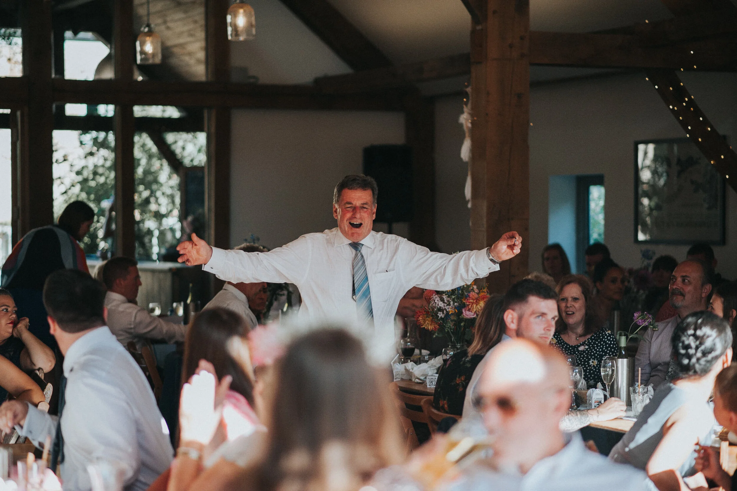 Man in white shirt and tie standing with arms outstretched, smiling at a celebration or gathering in a decorated rustic indoor venue.