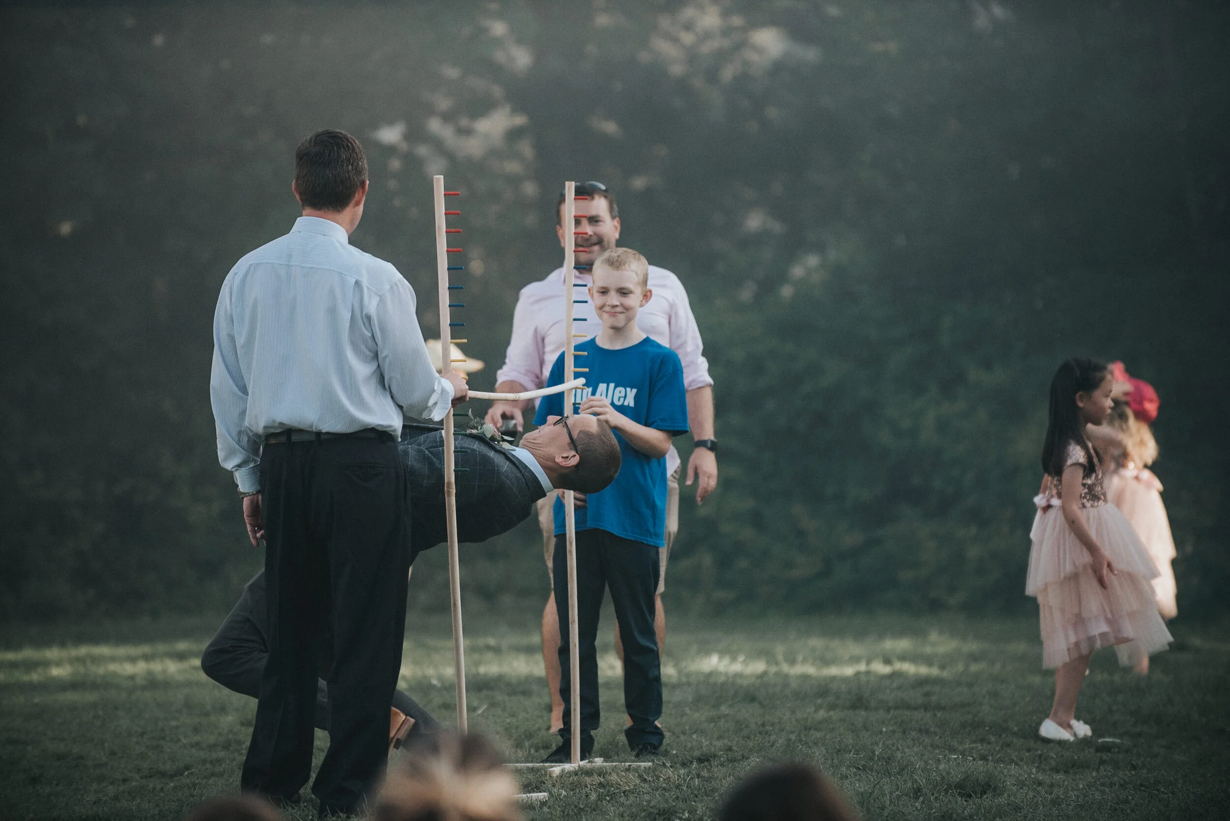 Group of children playing limbo outdoors on grass with trees in background, two girls wearing dresses to the right, adults observing in the background.