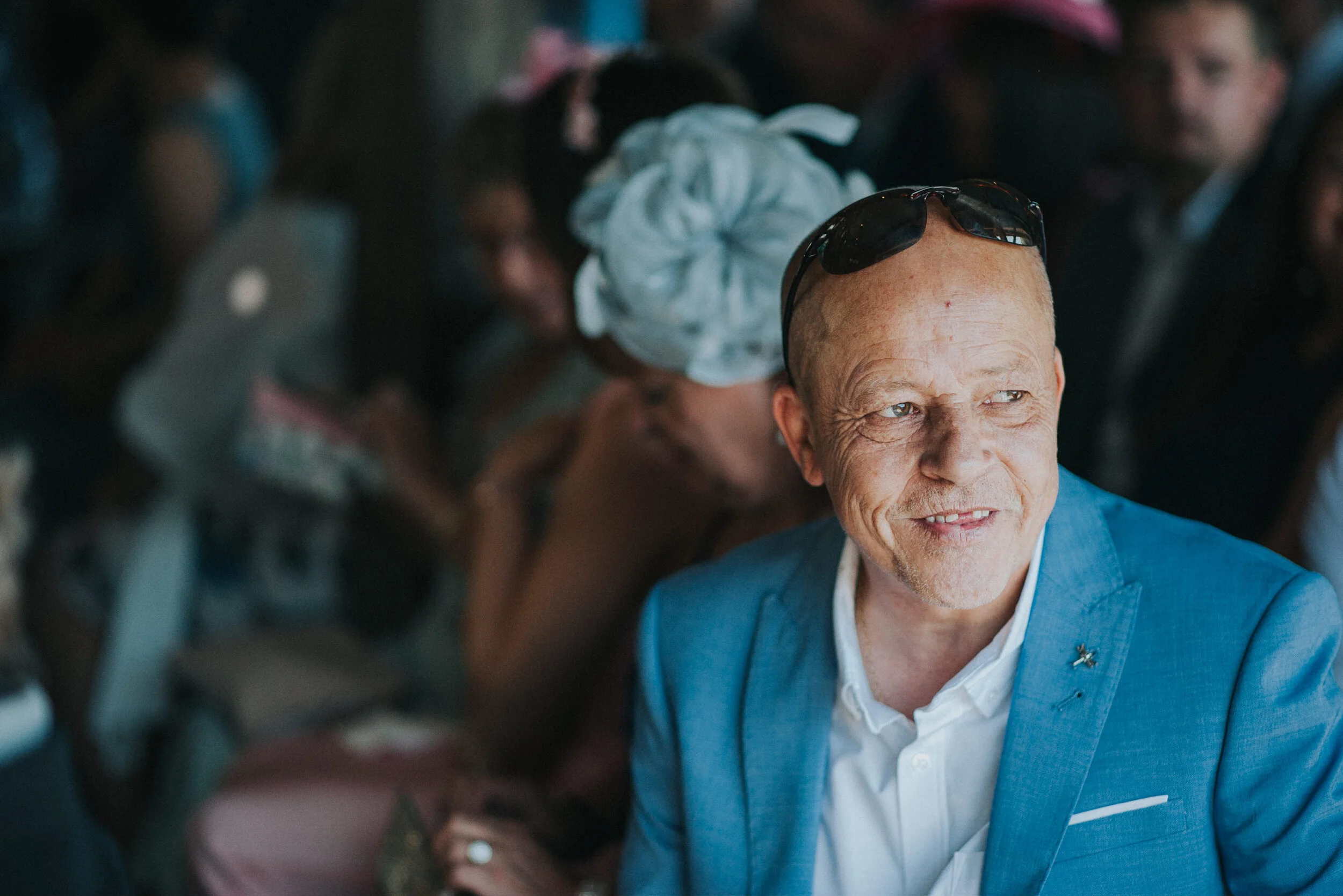 A smiling man with a shaved head wearing sunglasses on his forehead, dressed in a bright blue blazer and white shirt, sitting at an event with blurred people in the background.