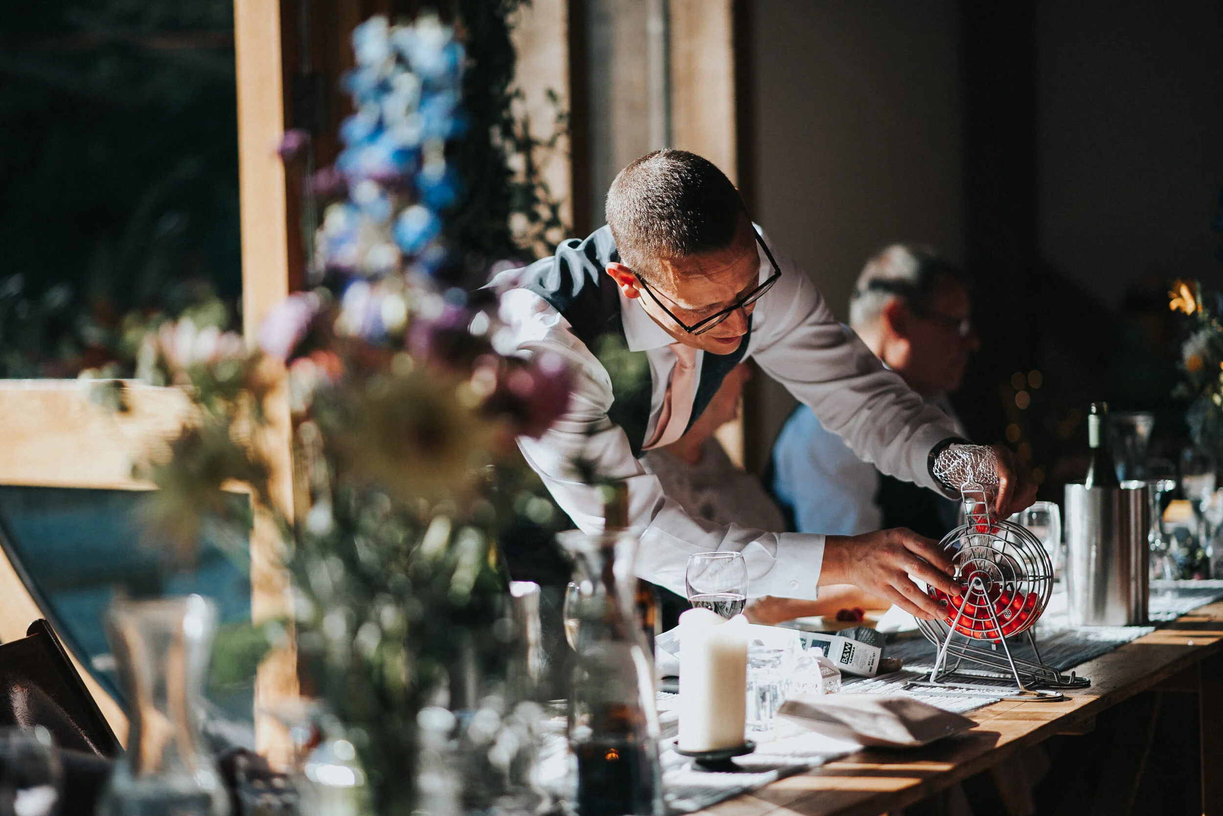 A man with glasses in a white shirt and dark vest picks a raffle ticket from a stand at a wooden table during a celebration or event, with other people and decorations visible in the background.