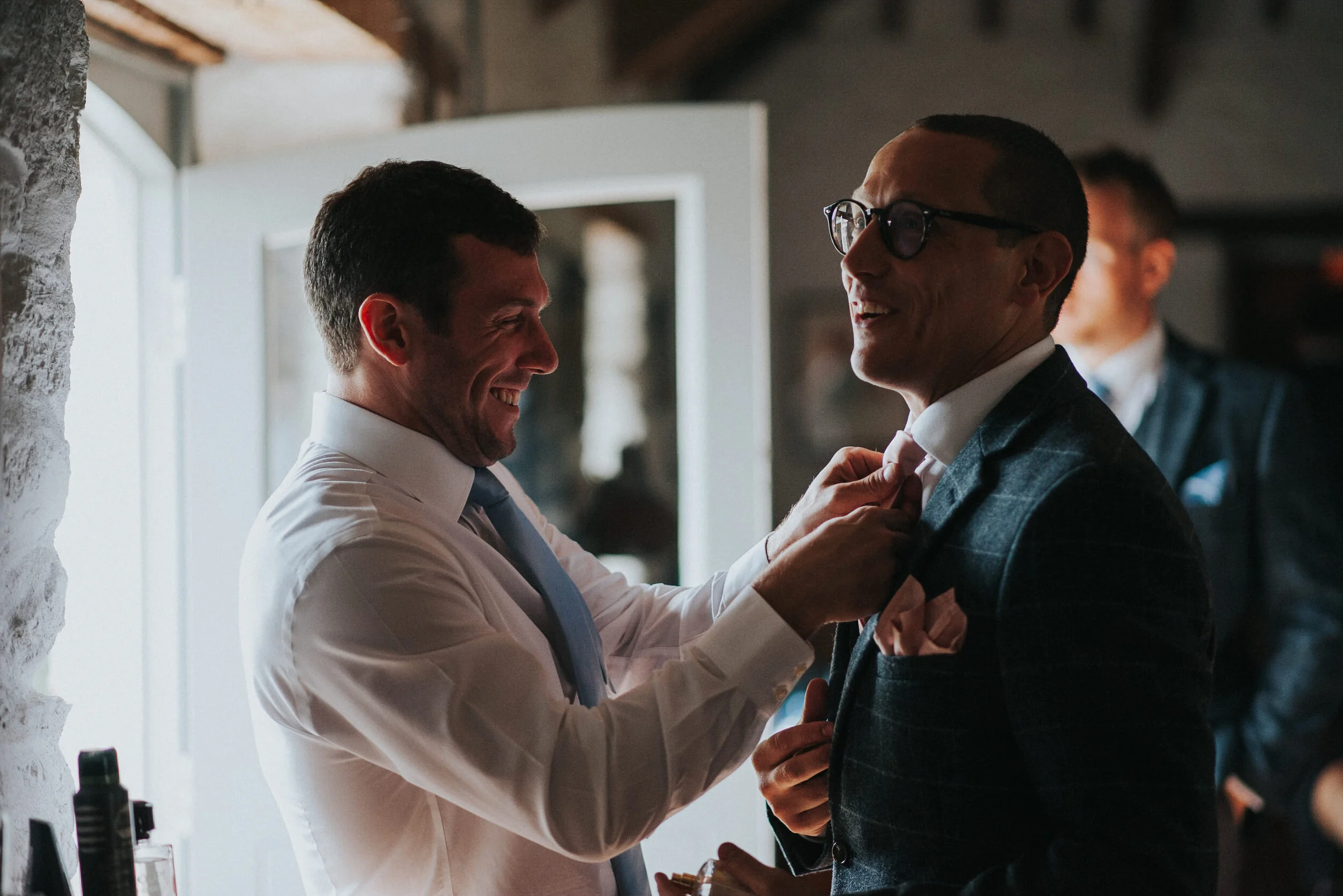 Two men in suits smiling and interacting, one adjusting the other's tie, indoors with natural light.