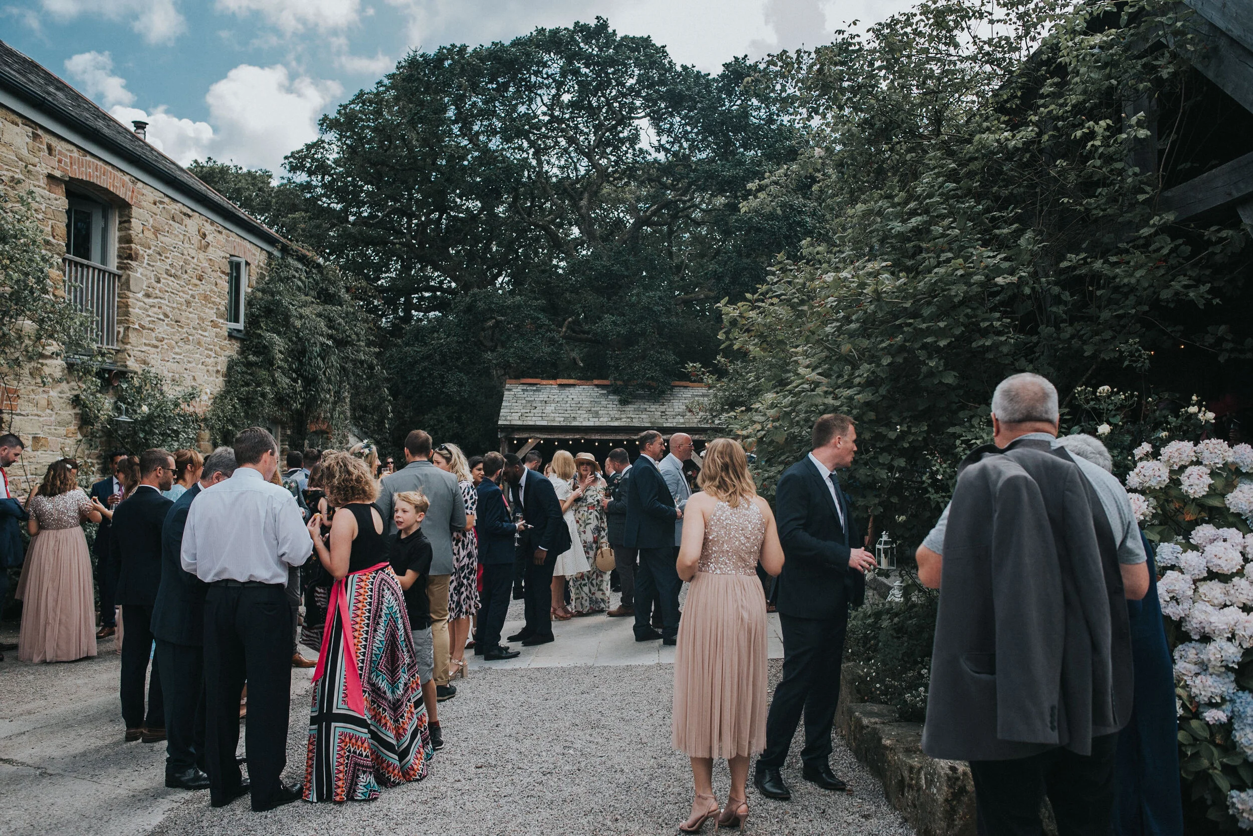 Nancarrow Farm Wedding Photography - People gathering outdoors during a social event, standing on gravel, with a stone building and lush greenery in the background.