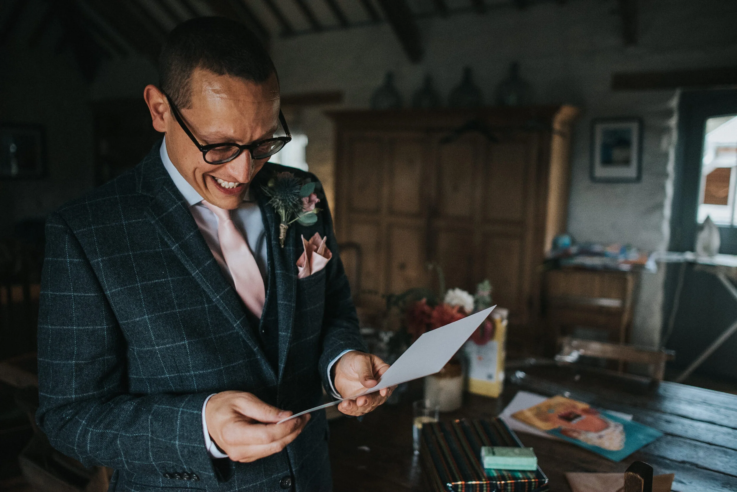 A man dressed in a plaid suit with a pink tie and boutonnière, smiling and looking at a piece of paper inside a rustic room with wooden furniture and decorations.