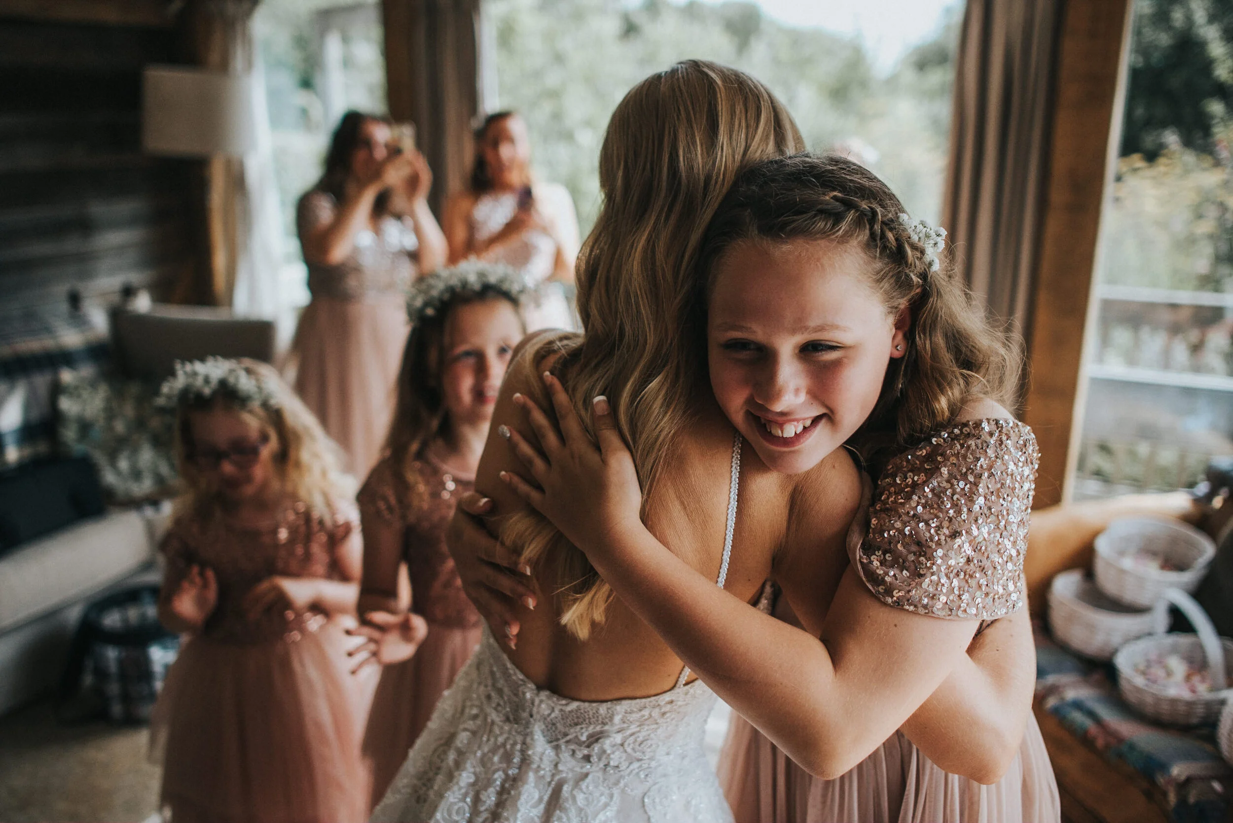 A young girl in a sequined dress hugs a woman in a white wedding gown, smiling, with three other girls in pink dresses and flower crowns in the background.