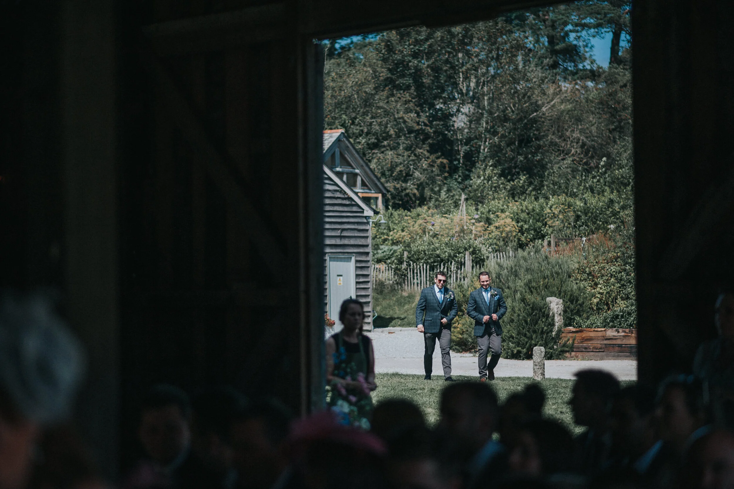 Two men dressed in suits walking outdoors in a garden area during daytime, viewed through an open barn door with a crowd inside, and trees and a wooden building in the background.