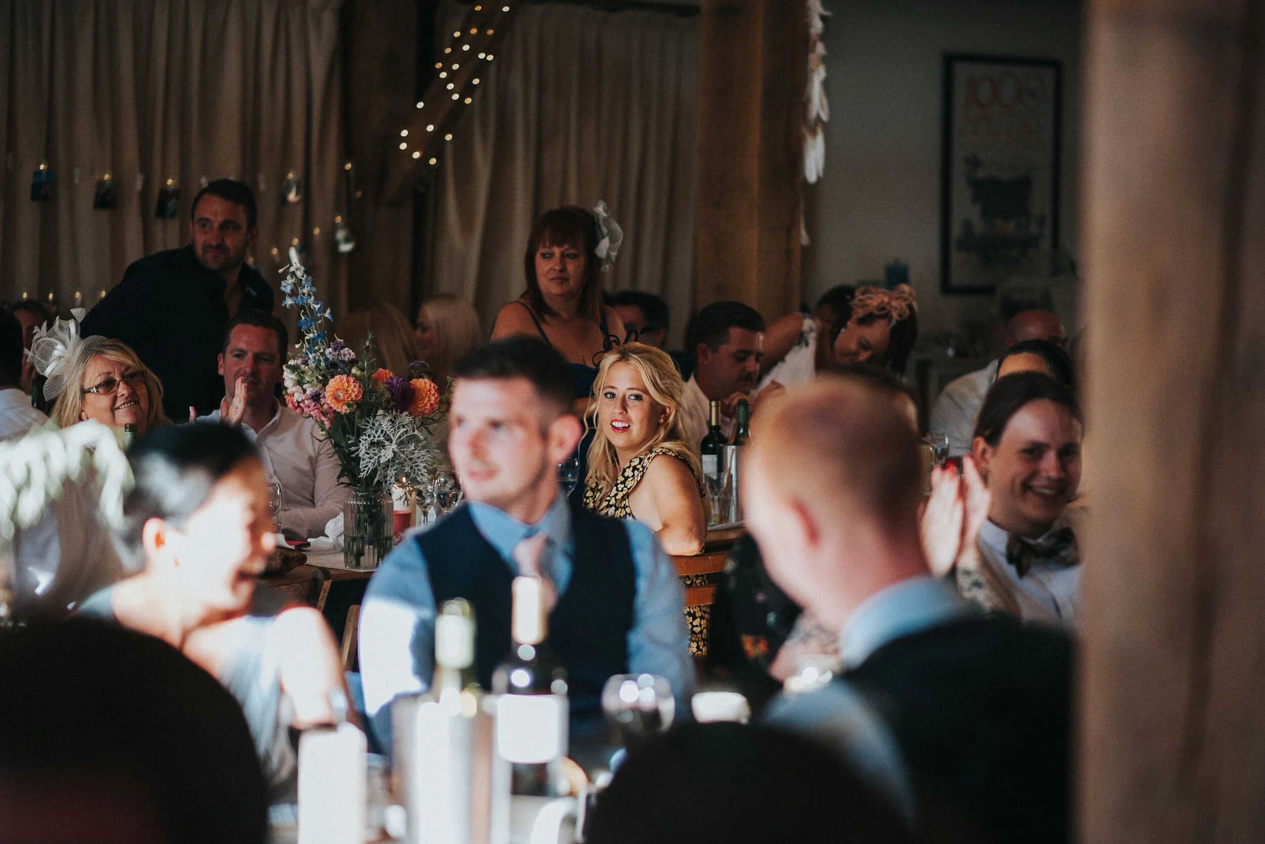 People at a wedding reception or celebration, sitting around tables with flowers, bottles, and glasses, some are laughing and smiling, with warm background lighting and decorations.