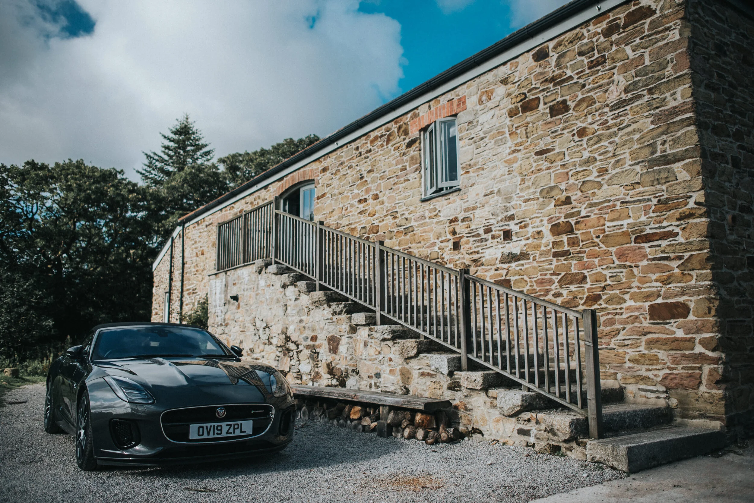 A black Jaguar car parked next to a stone building with a staircase and a railing, surrounded by trees under a partly cloudy sky.