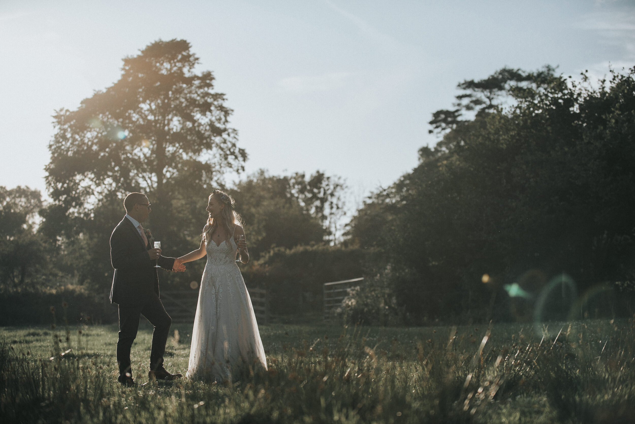 A couple in wedding attire holding hands outdoors during sunset. The man is in a suit and glasses, the woman in a white wedding dress, standing on a grassy field with trees in the background.
