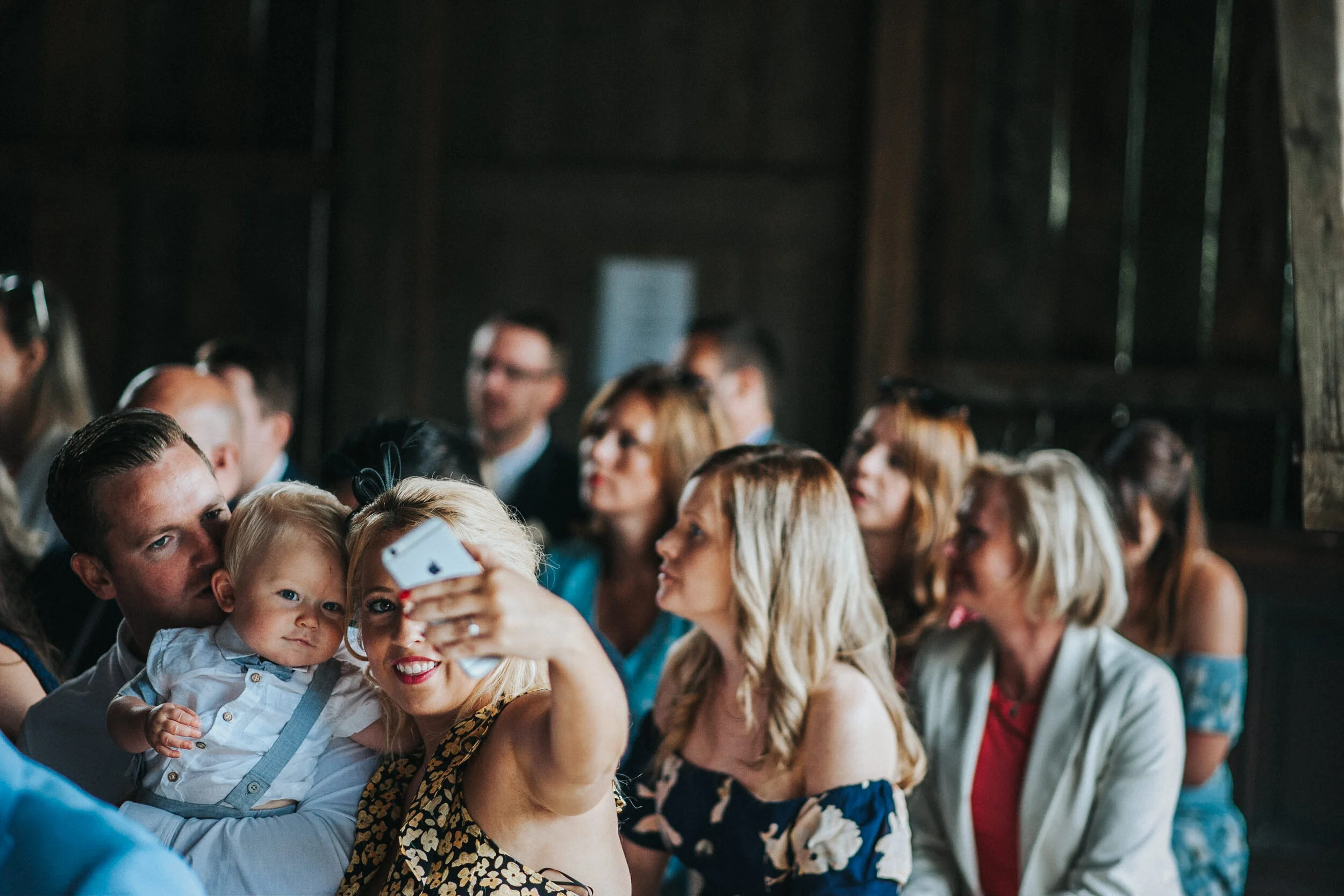 Group of people seated at a social gathering, with woman in the foreground taking a selfie, holding a young boy in her lap, both looking at the camera, in a rustic indoor setting.