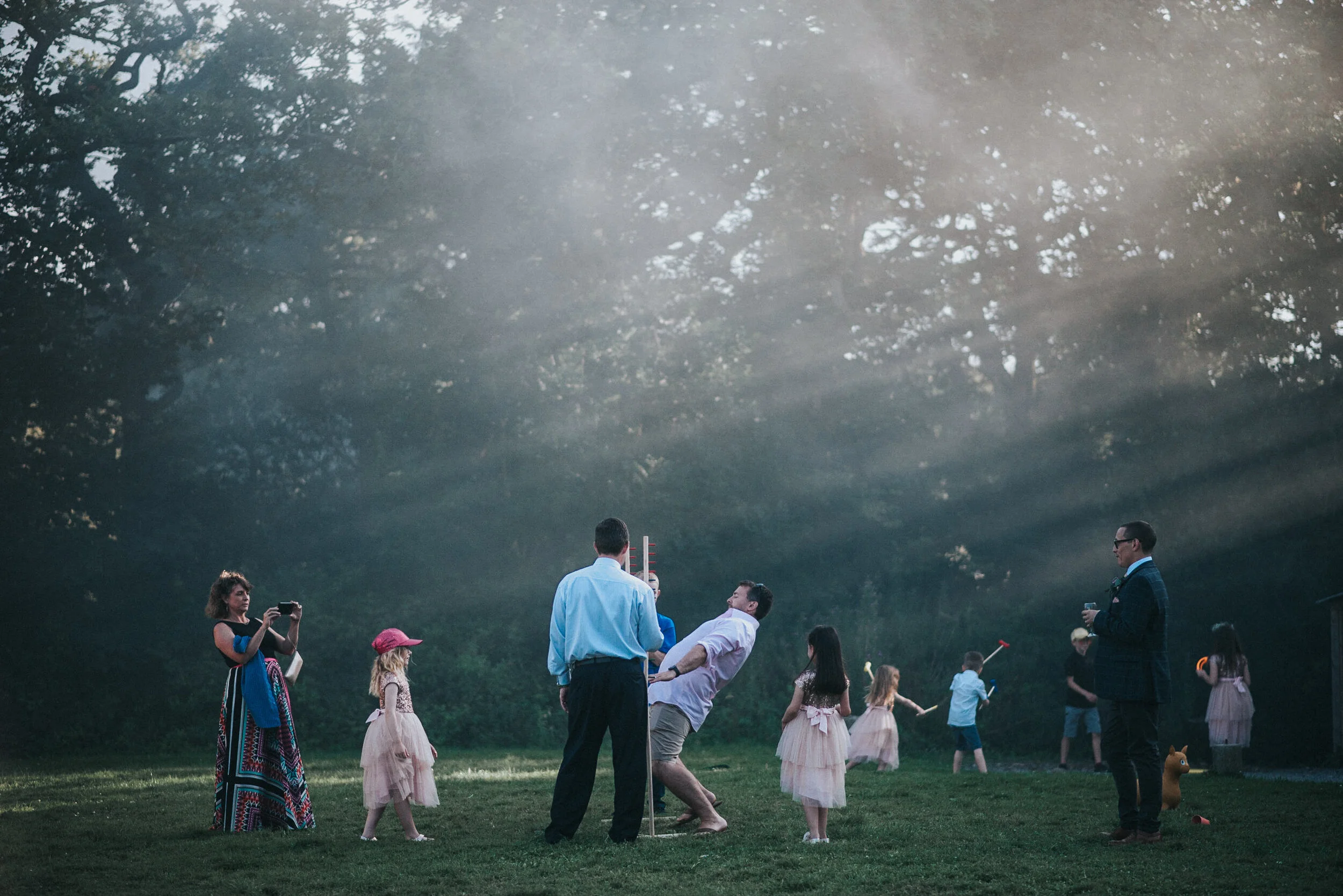 A group of children and adults on a grassy field with trees in the background plays and takes photos, some holding toys and balloons, with sunlight streaming through the trees.