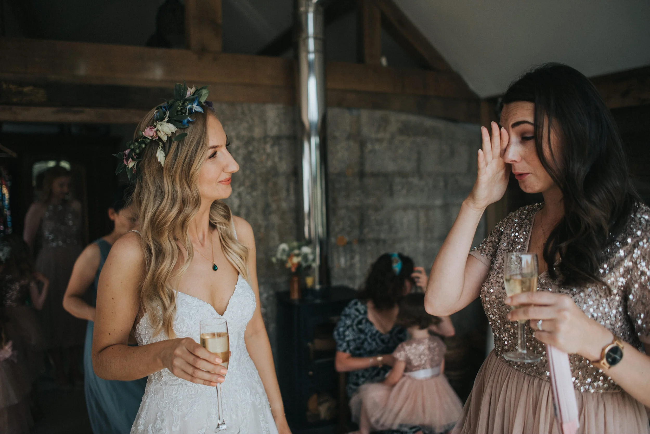 Two women at a wedding reception, one in a white wedding dress with a floral crown, holding a glass of champagne, discussing with another woman in a sparkly dress, also holding a glass of champagne, with other guests and children in dresses in the ba