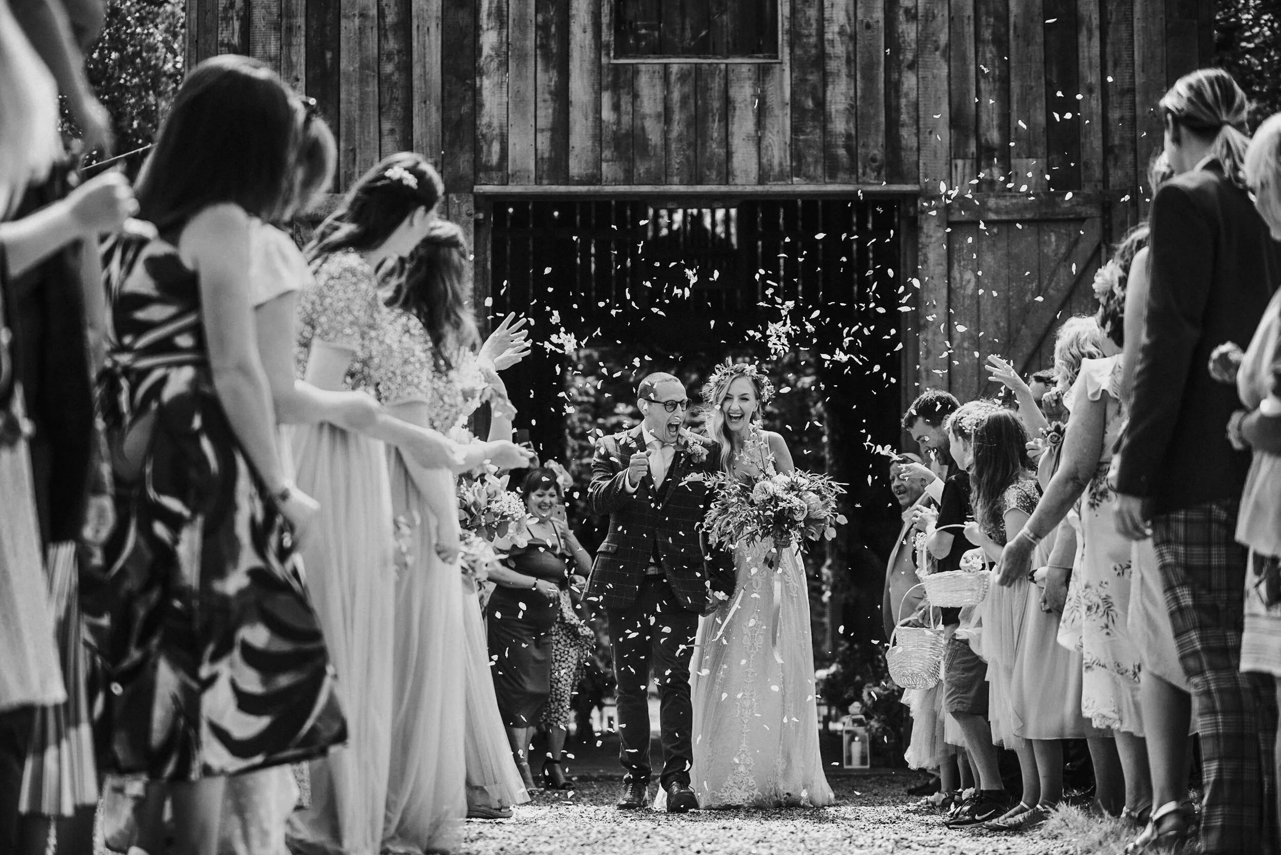 A black and white photo of a bride and groom walking through a crowd of wedding guests throwing confetti at them, outdoors near a rustic wooden barn.