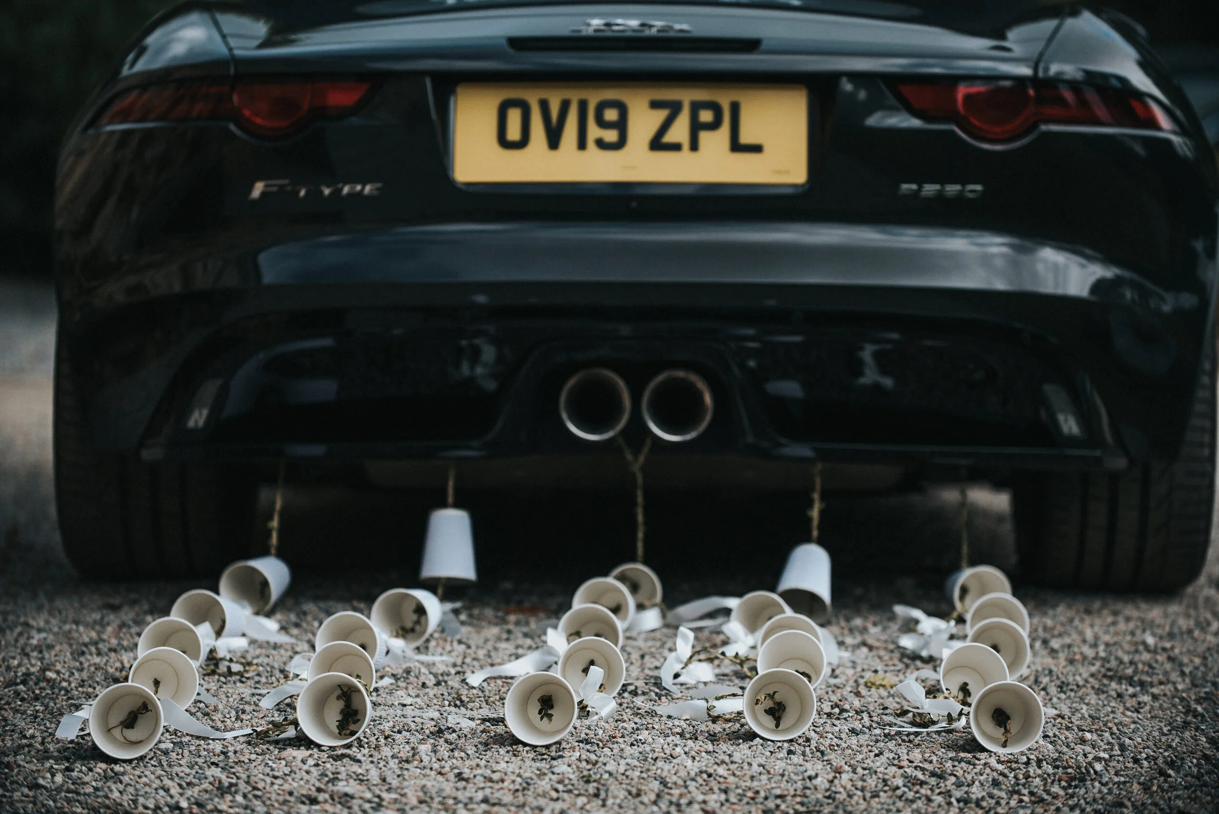 Black sports car with a yellow license plate and a dual exhaust pipe, surrounded by empty white coffee cups lying on the ground.