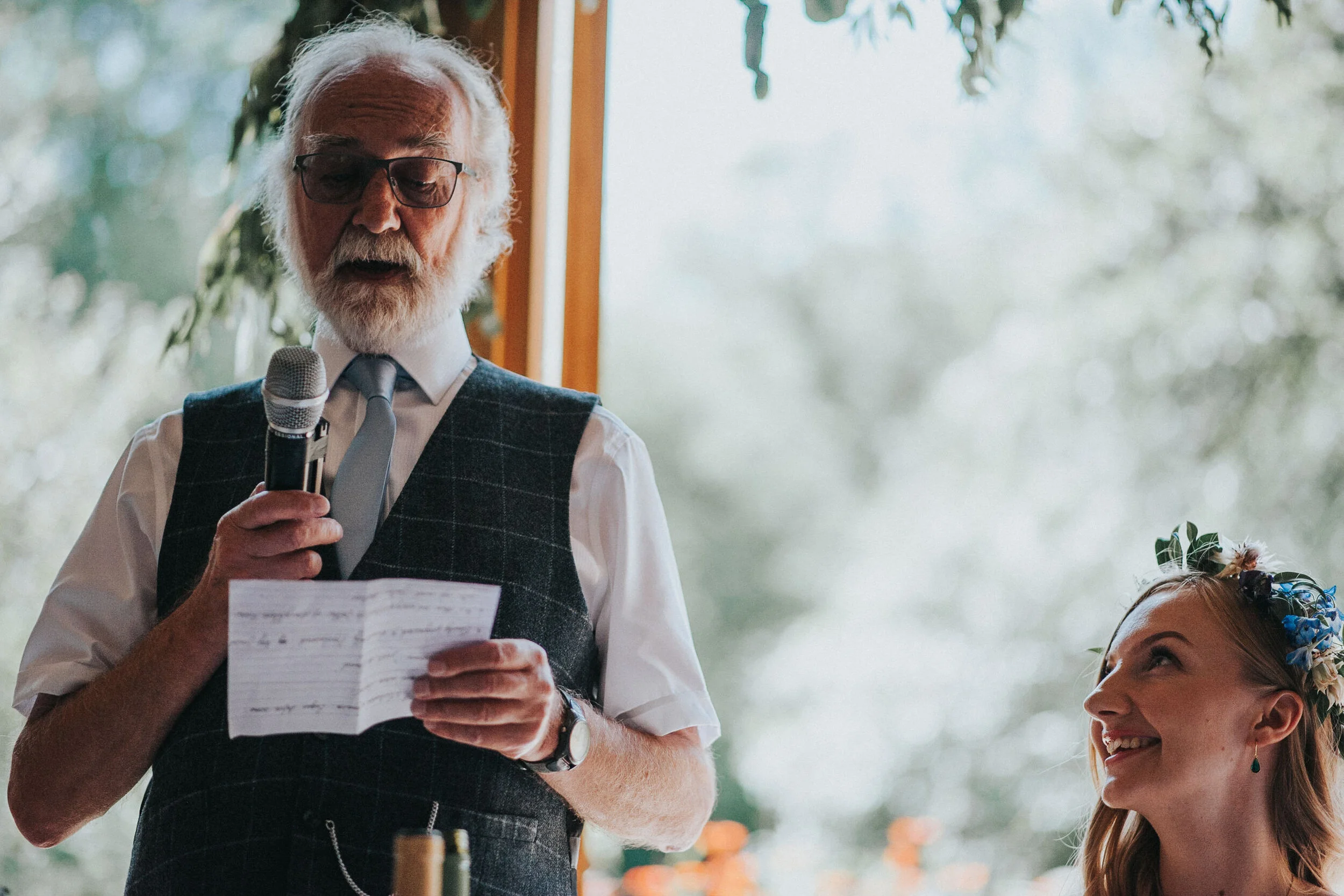 An elderly man with white hair and beard, wearing glasses, a white shirt, grey vest, and tie, is holding a microphone and reading from a piece of paper. A young woman with long hair, floral headband, and earrings is looking up at him, smiling.