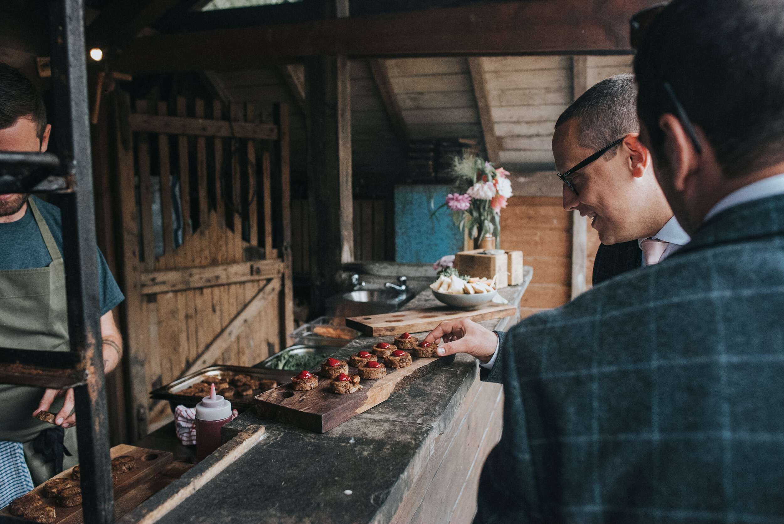 Two men in suits at a rustic food stand, selecting small appetizers with a pier, a bowl of chips, and flowers in the background.