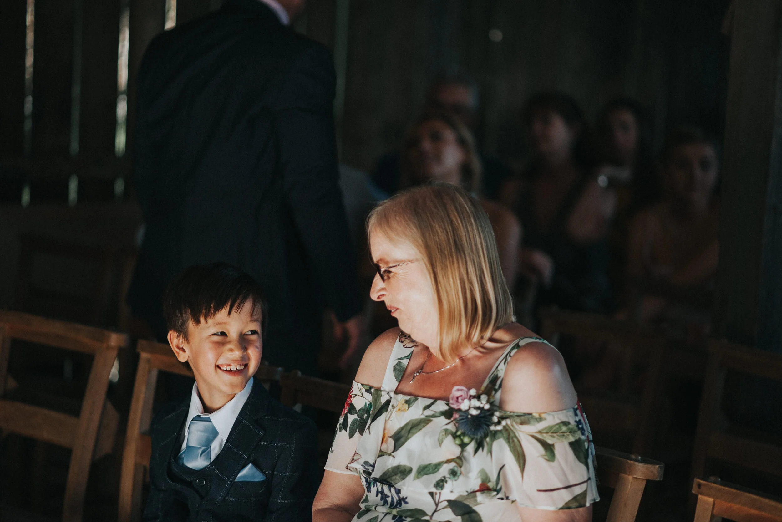 A young boy and an older woman, likely a grandmother, sitting and laughing together at a social event with other people in the background.