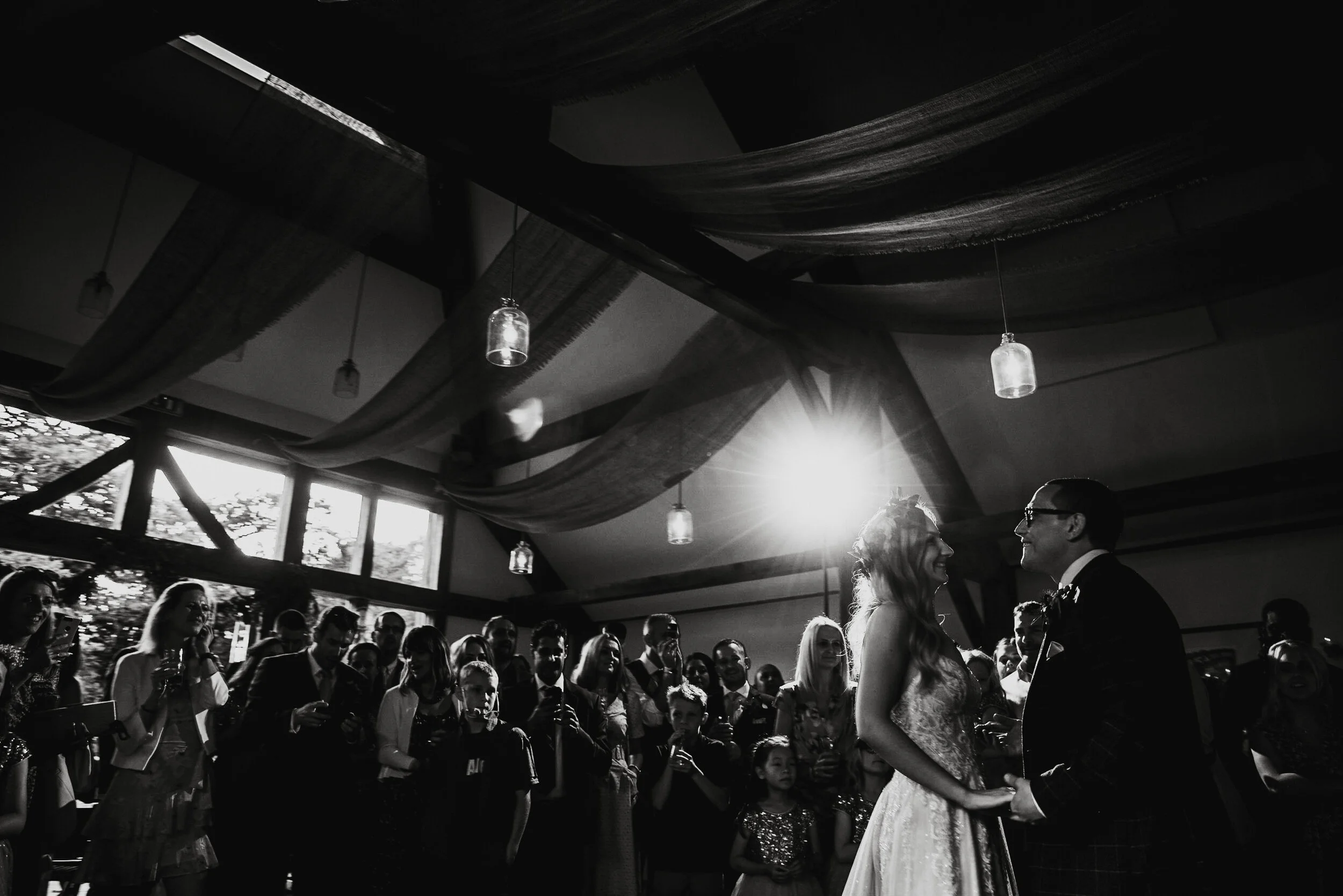 Black and white photograph of a wedding ceremony with the bride and groom holding hands and smiling at each other, surrounded by wedding guests inside a decorated venue with hanging lights and draped fabric.