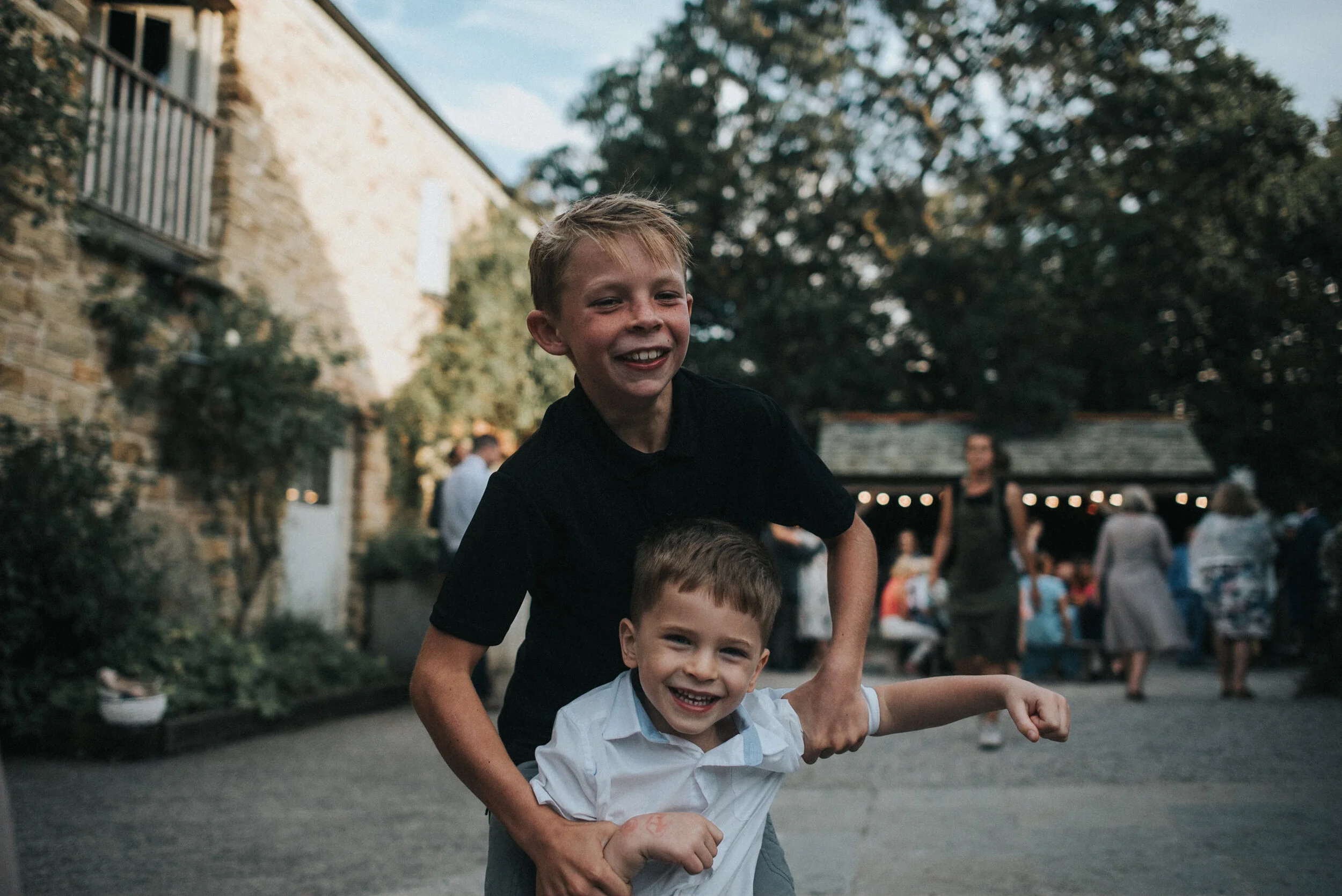 Two boys smiling and playfully wrestling outdoors at a gathering or party. The older boy has blonde hair and is wearing a black shirt, and the younger boy has brown hair and is wearing a white shirt. In the background, other people are socializing ne