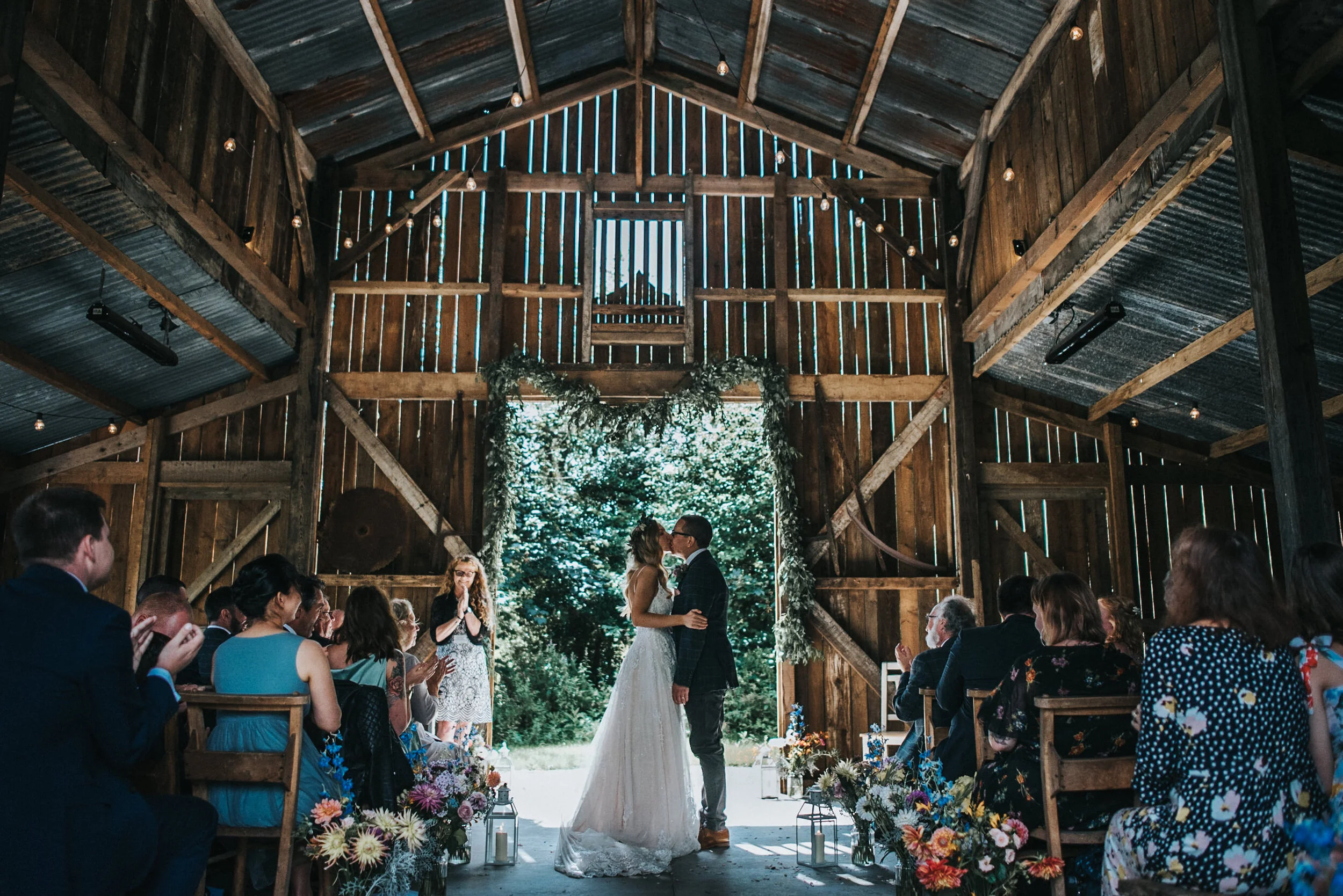 A wedding ceremony inside a rustic barn, with a bride and groom kissing in front of an audience of seated guests, decorated with flowers and greenery.