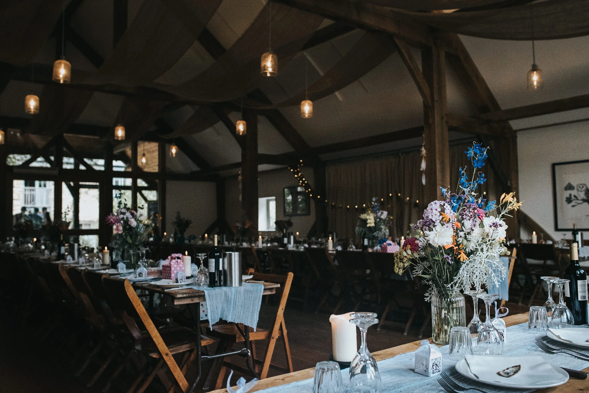 A decorated dining area with tables adorned with flowers, candles, and tableware inside a rustic venue with wooden beams and hanging lights.