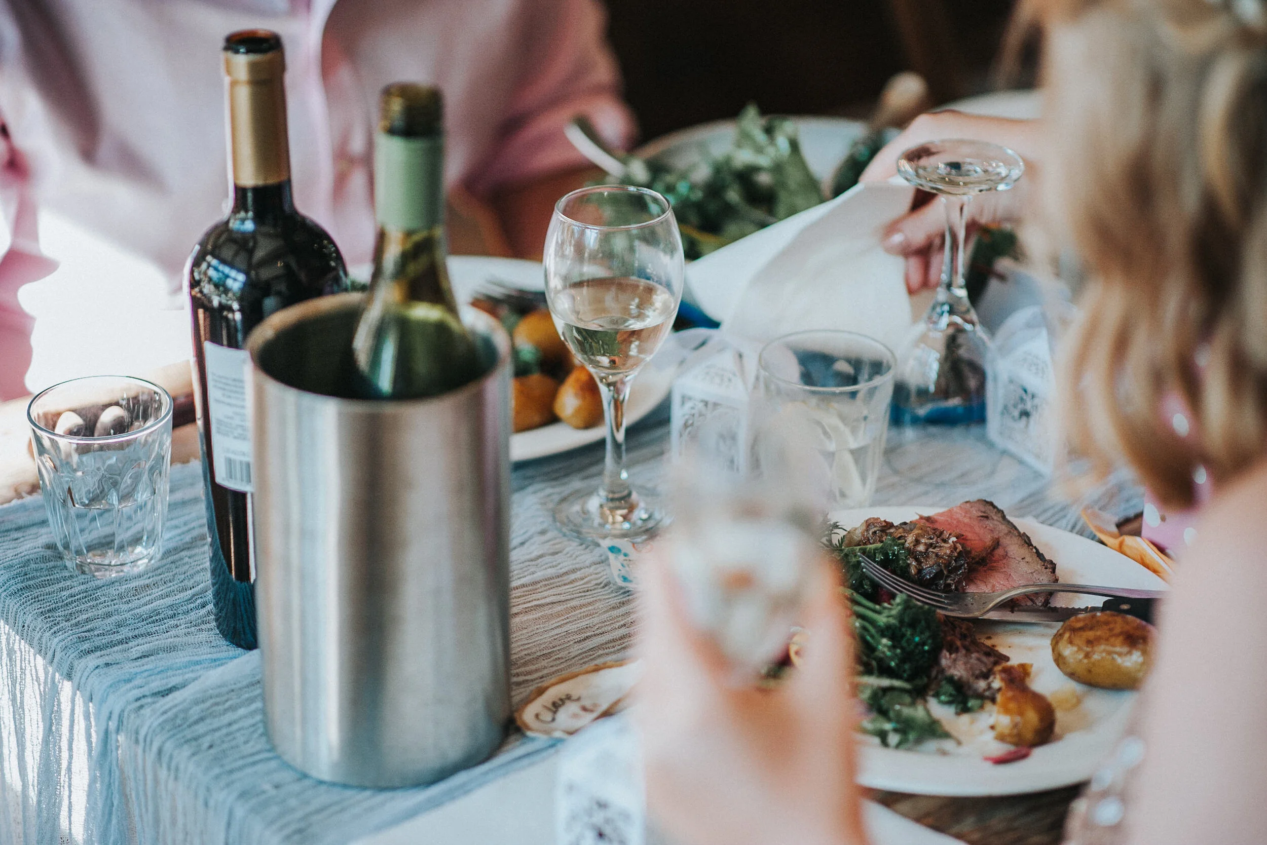 People dining at a table with wine bottles, glasses, a partially eaten plate of food, and a salad.